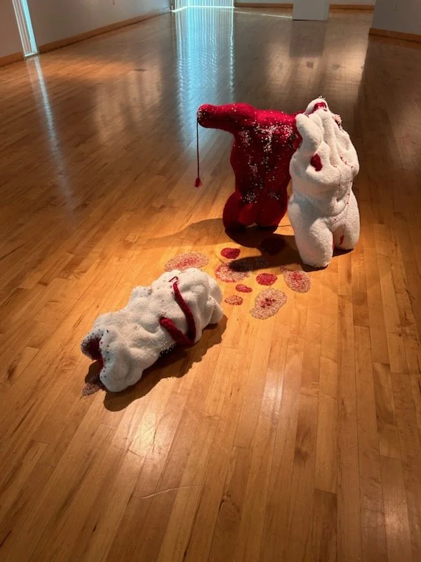 A white and red, torso sculpture that lies on a hardwood floor surrounded by scattered red and silver decorative pearls.