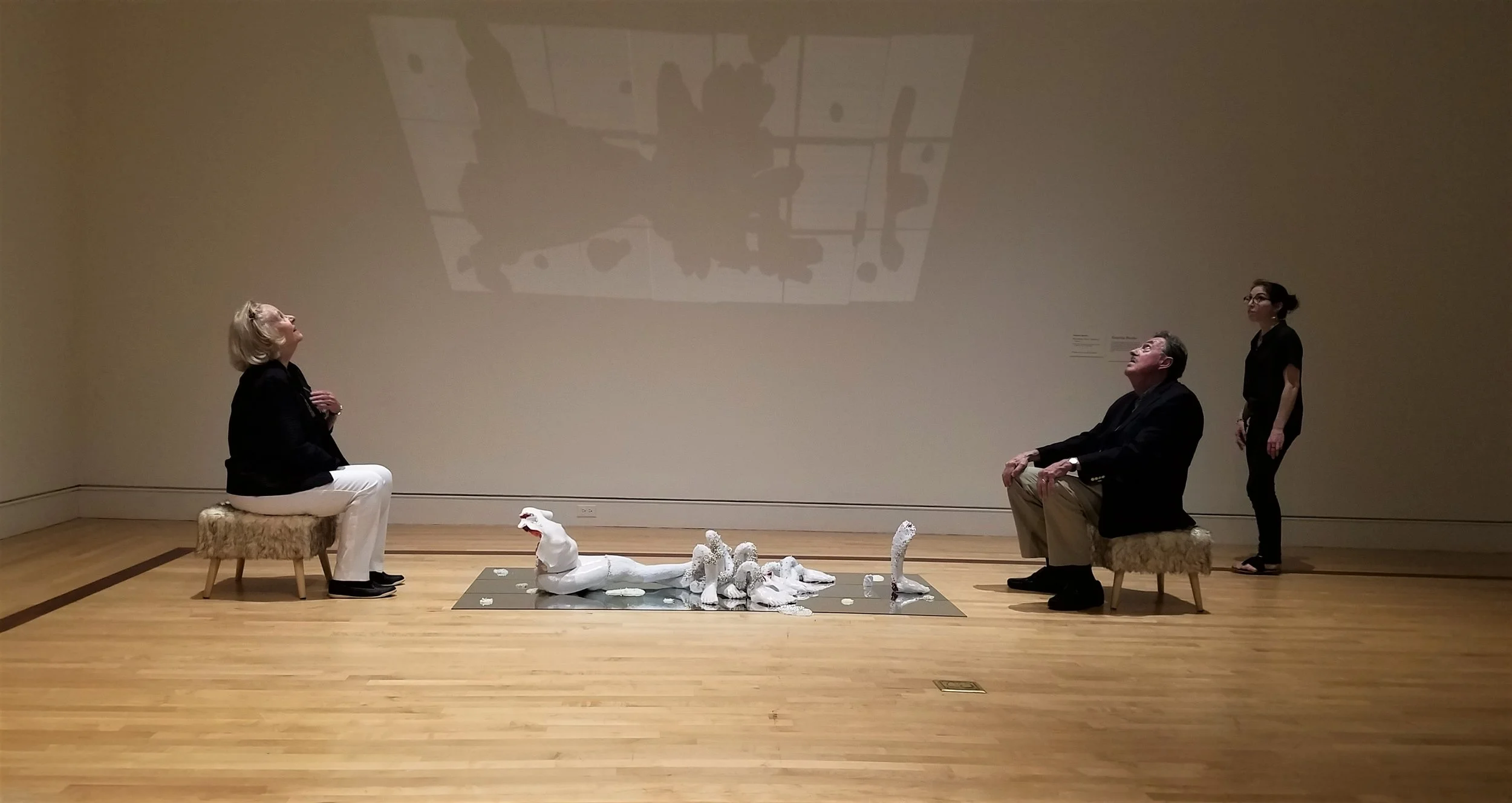 An art installation in a gallery depicting two people seated facing each other with a sculpture on the floor between them, and a woman standing behind a stool on the right. The shadows of multiple figures are cast on the wall behind them.