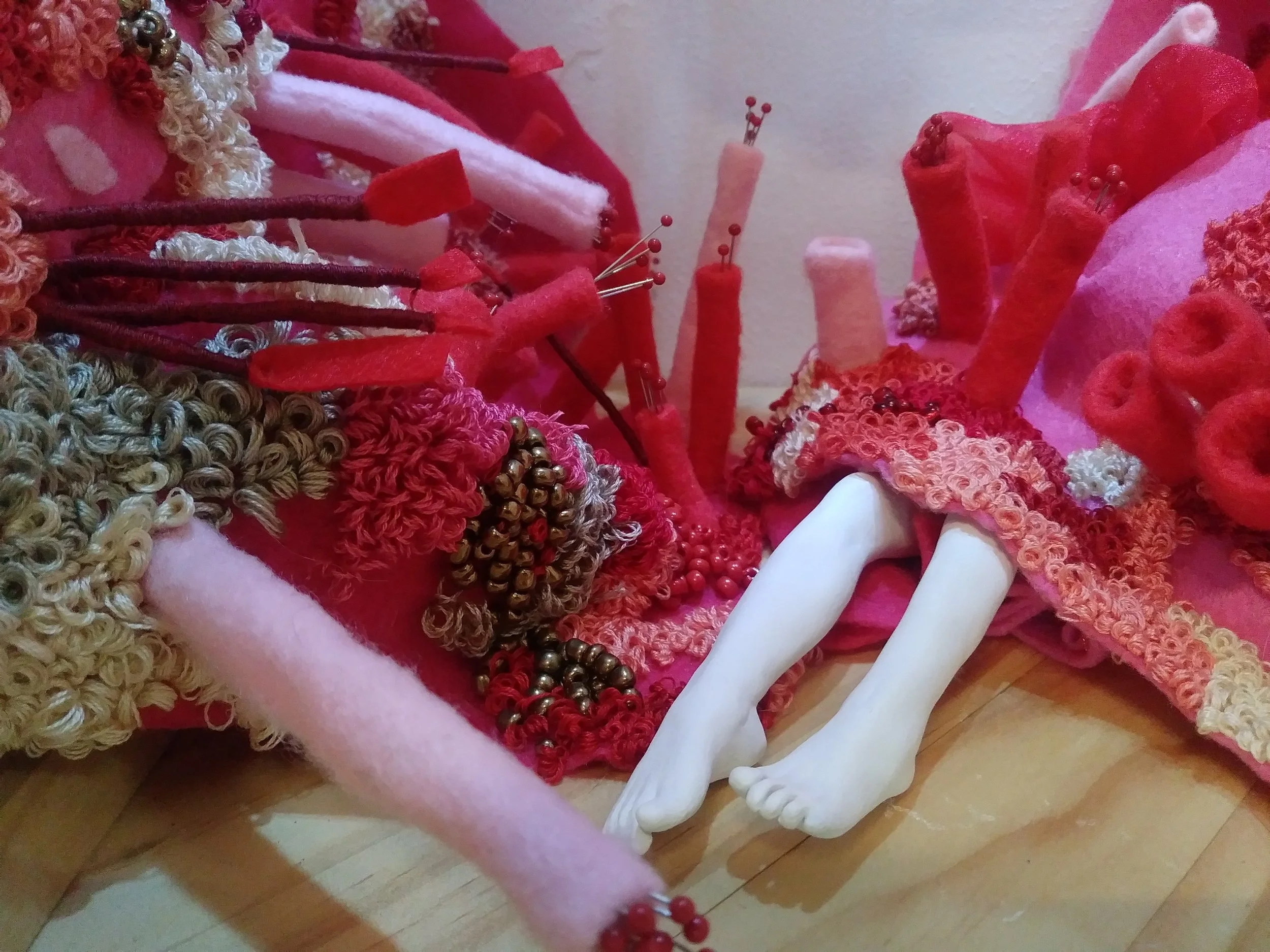 A detailed close-up image of various red, pink, and beige textiles and materials including yarn, felt, and beads, with some small parts resembling a mannequin or sculpture bones.