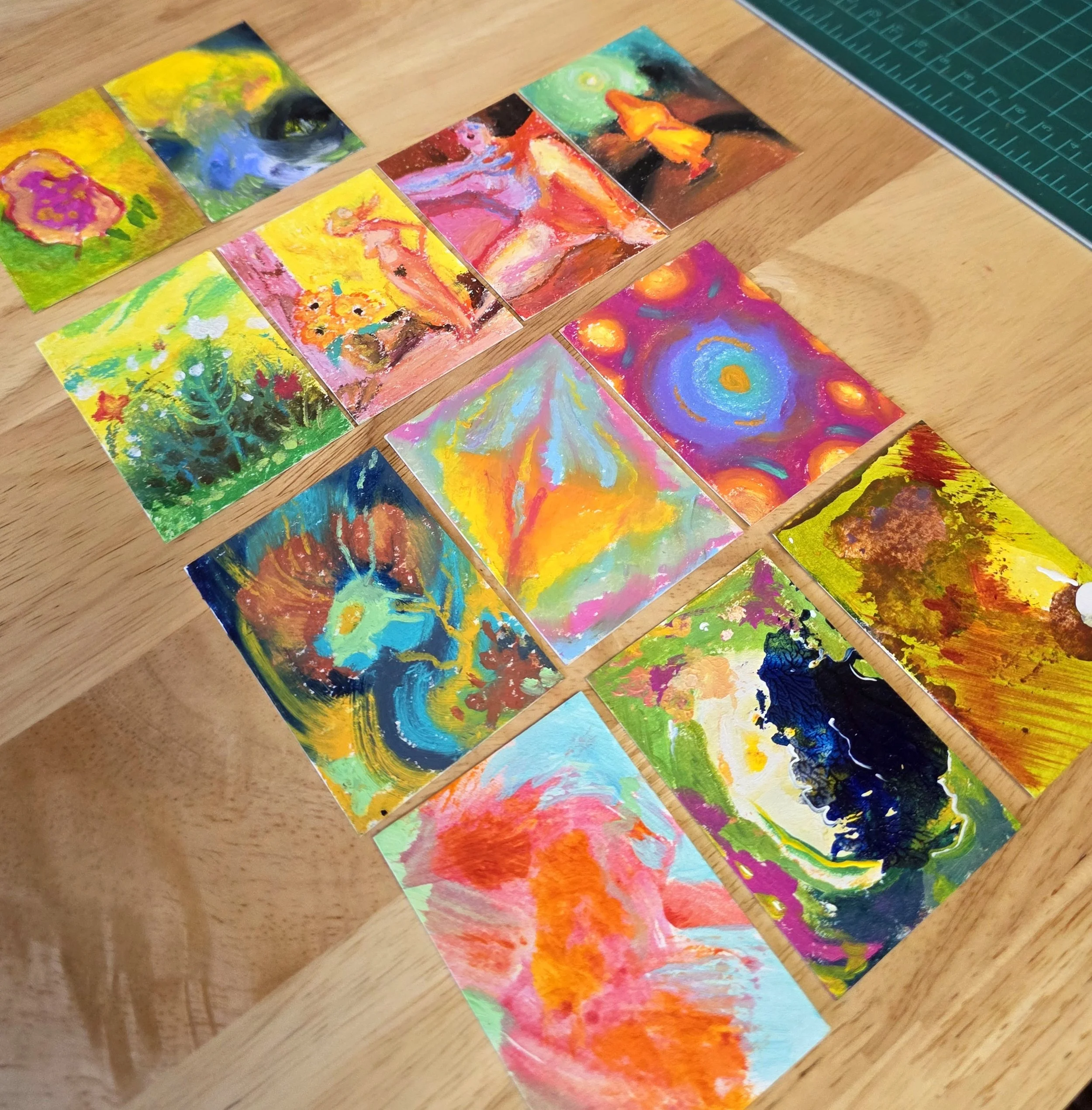 Colorful abstract paintings arranged on a wooden table with a green cutting mat in the corner.