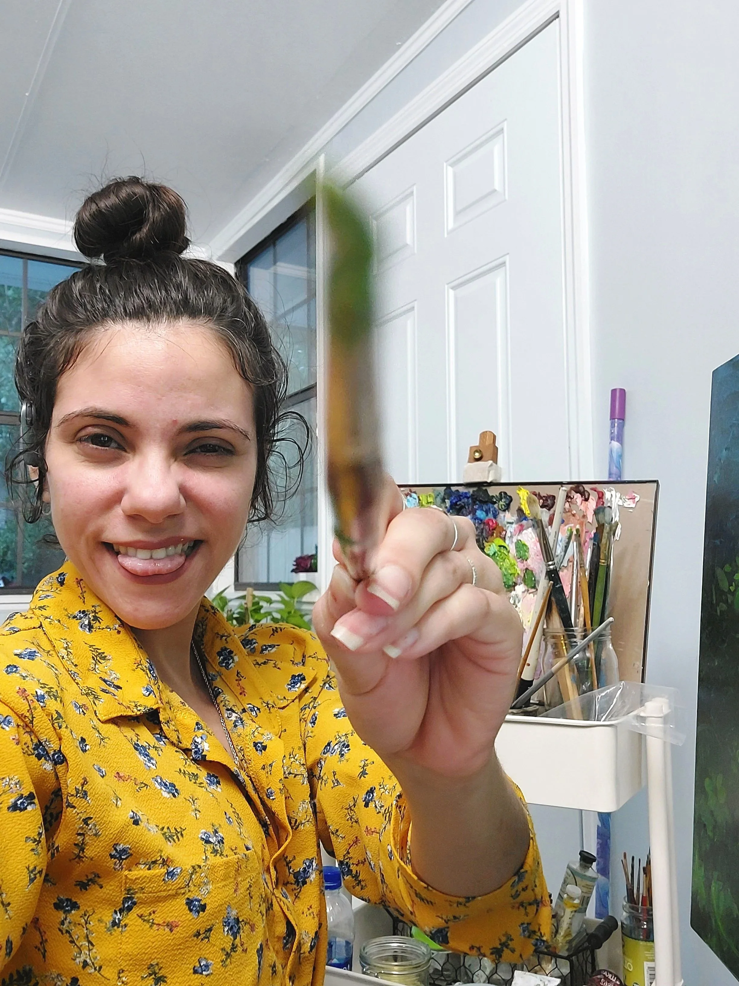 A woman with brown hair in a bun, wearing a yellow floral blouse, smiling and holding a blurred green and yellow brush in front of her, in an art studio or workspace with various supplies and paintings in the background.