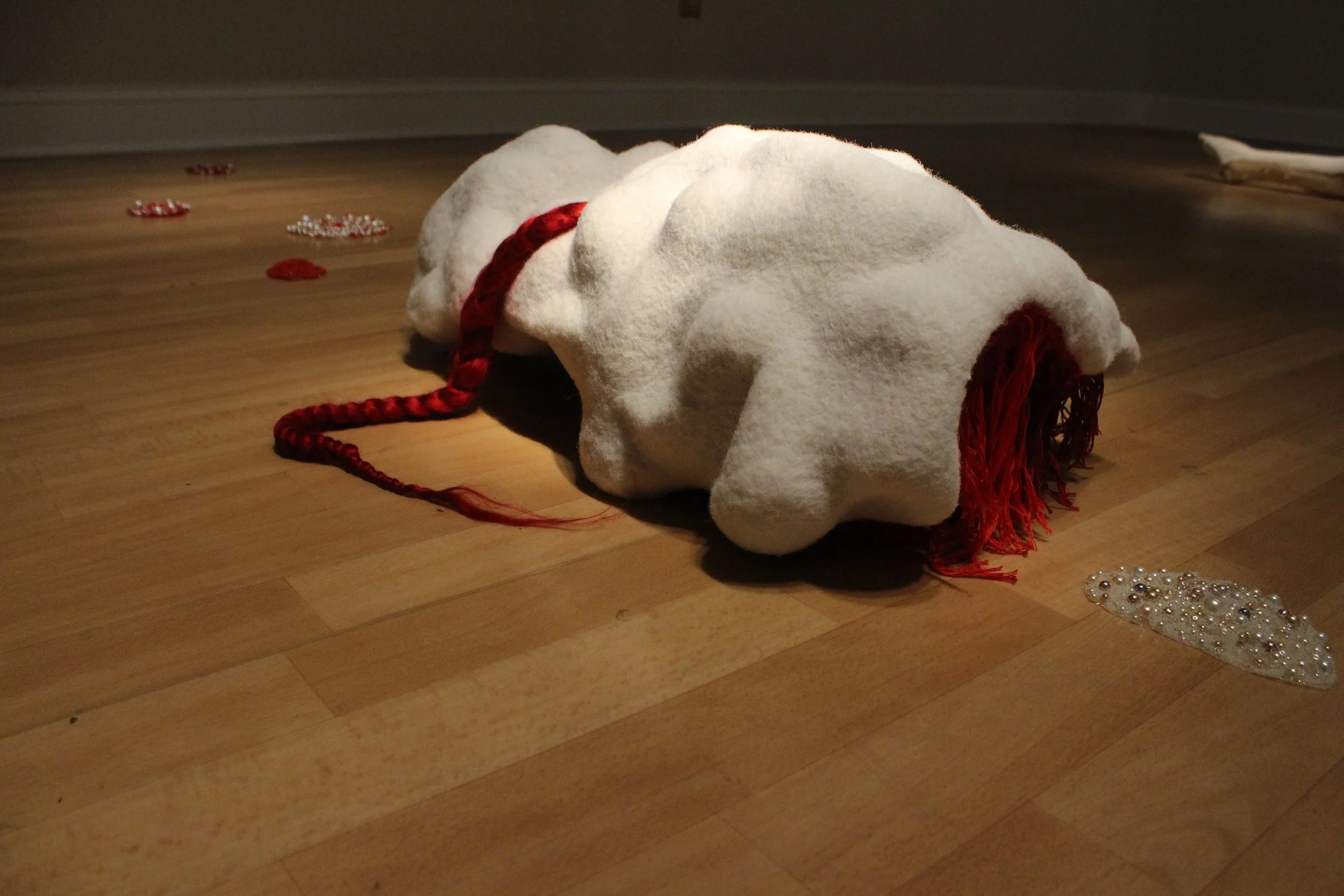 A white, shapeless, white fiber sculpture that lies on a hardwood floor surrounded by scattered red and silver decorative pearls.
