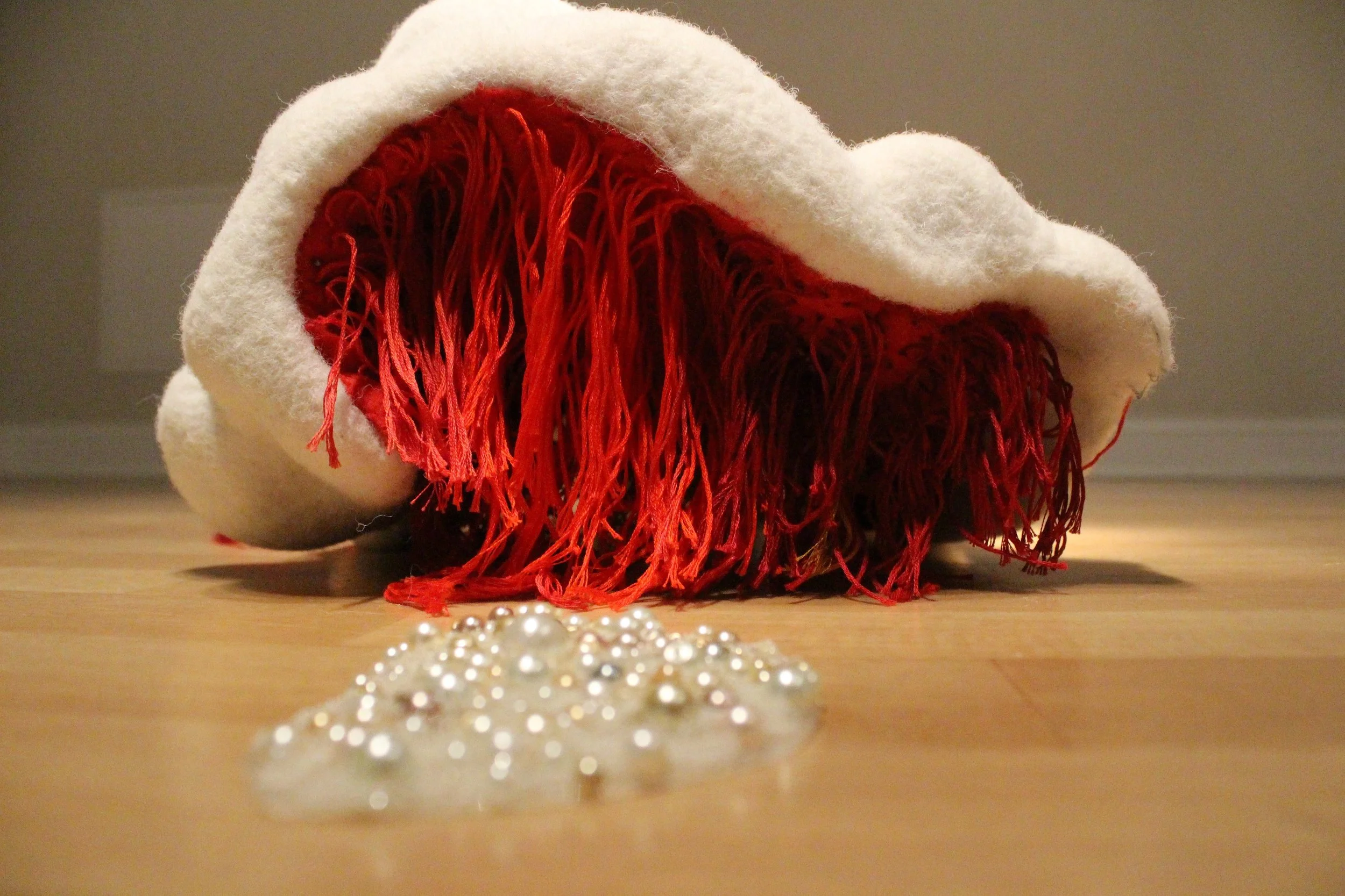 A white, shapeless, white fiber sculpture that lies on a hardwood floor surrounded by scattered red and silver decorative pearls.