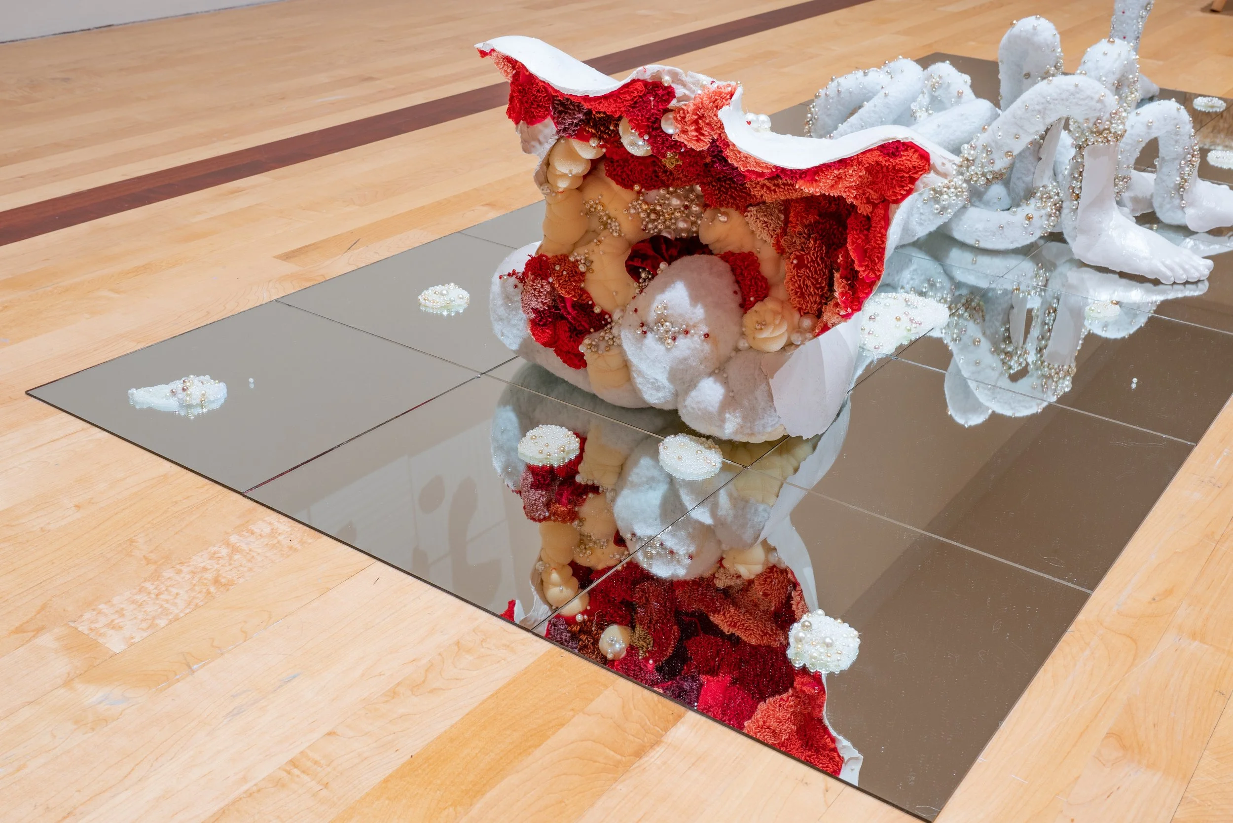 An art sculpture  with a flowing red and white fabric adorned with beads and decorations, placed on a reflective surface.