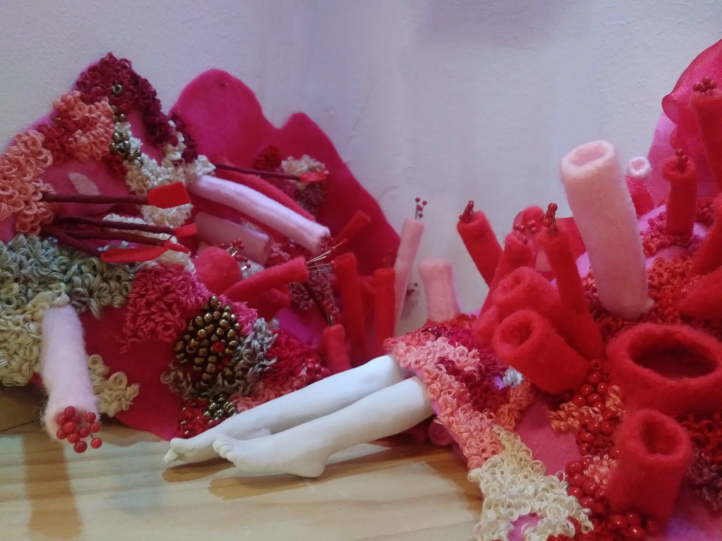 A collection of handmade fabric flowers in red, pink, and cream, with some adorned with beads and wire, arranged on a wooden surface against a plain wall.