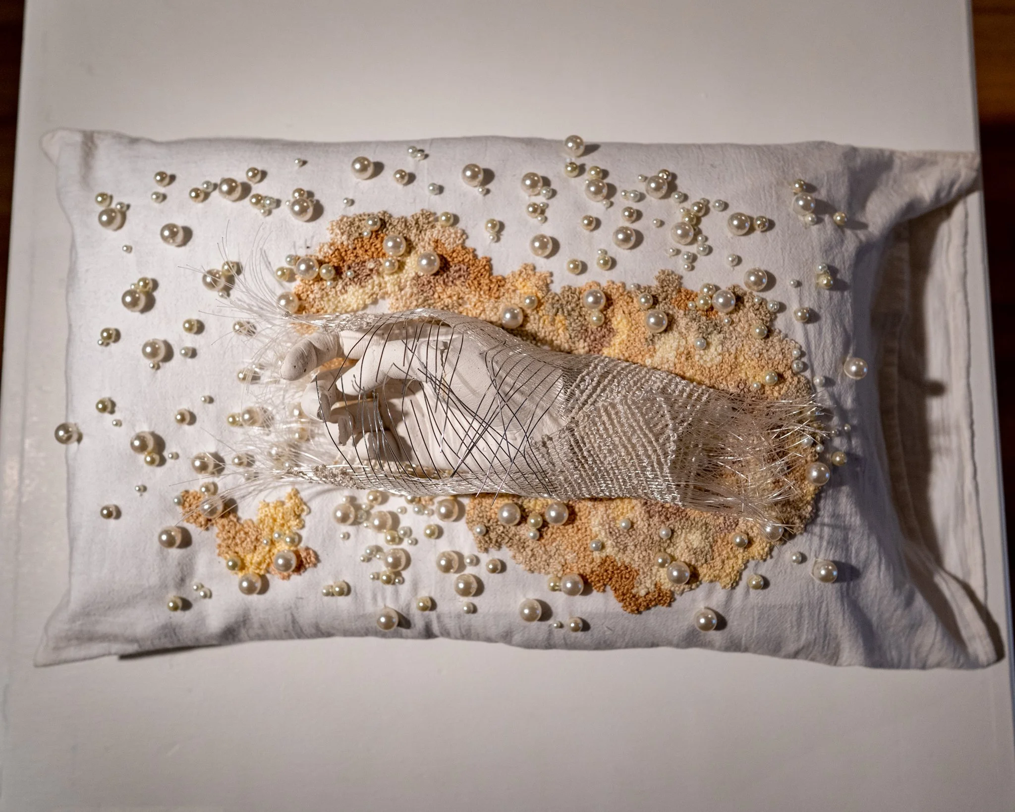 A white pillow decorated with scattered white pearls, tan, beige, and white textured beads, some metallic silver accents, and what appears to be a decorative net or veil underneath.