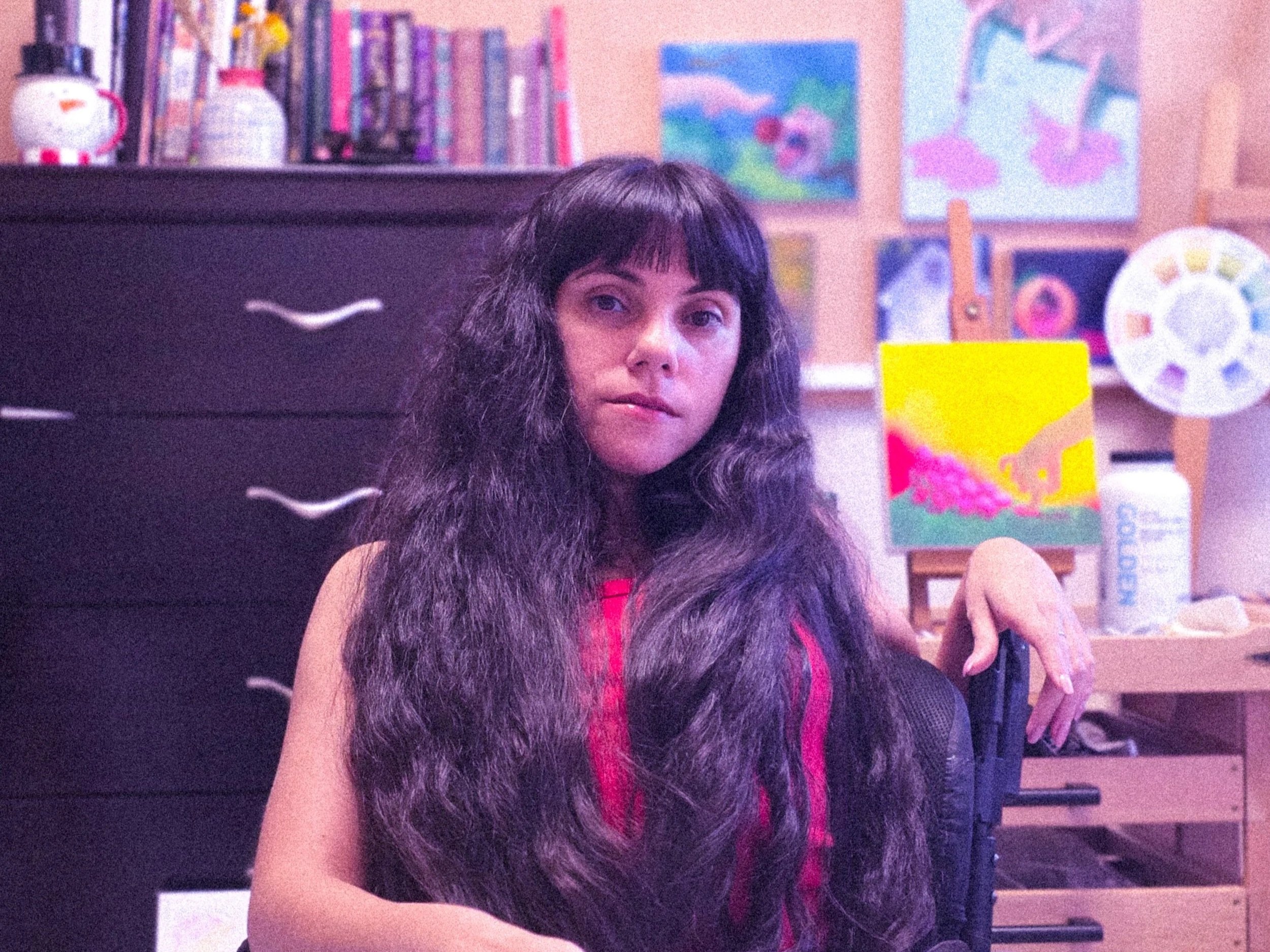A woman with long curly hair and bangs looking at the camera, sitting in a room decorated with colorful paintings and books, with a black speaker in front of her.