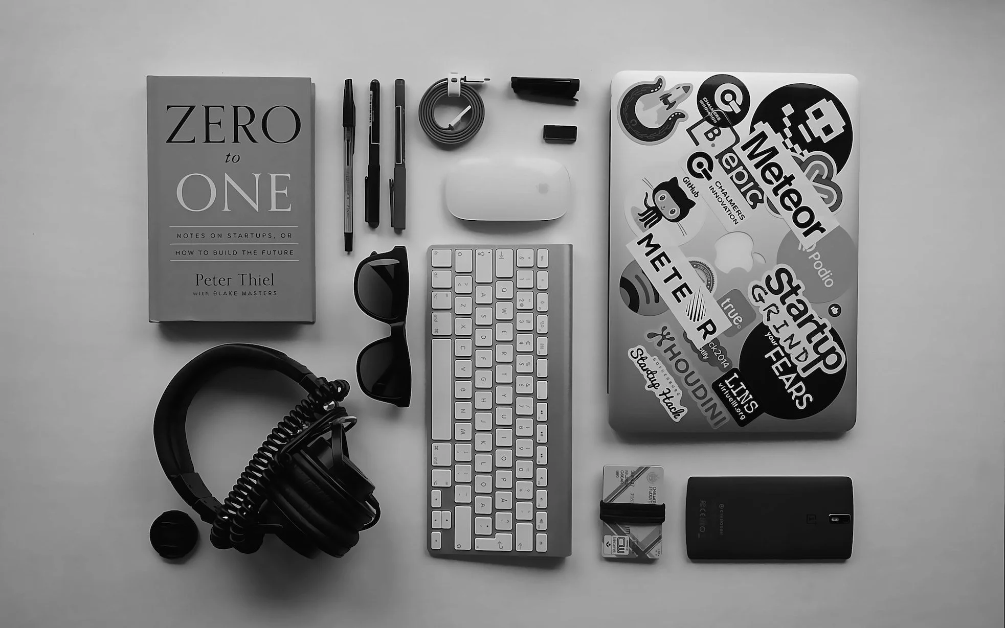 A flat lay of various tech gadgets, a book, and office supplies on a white surface. Items include headphones, sunglasses, a wireless keyboard, a smartphone, a laptop covered with stickers, a Wi-Fi router, a pack of memory cards, a notebook titled 'Zero to One', pens, a charging cable, and a small black plastic object.