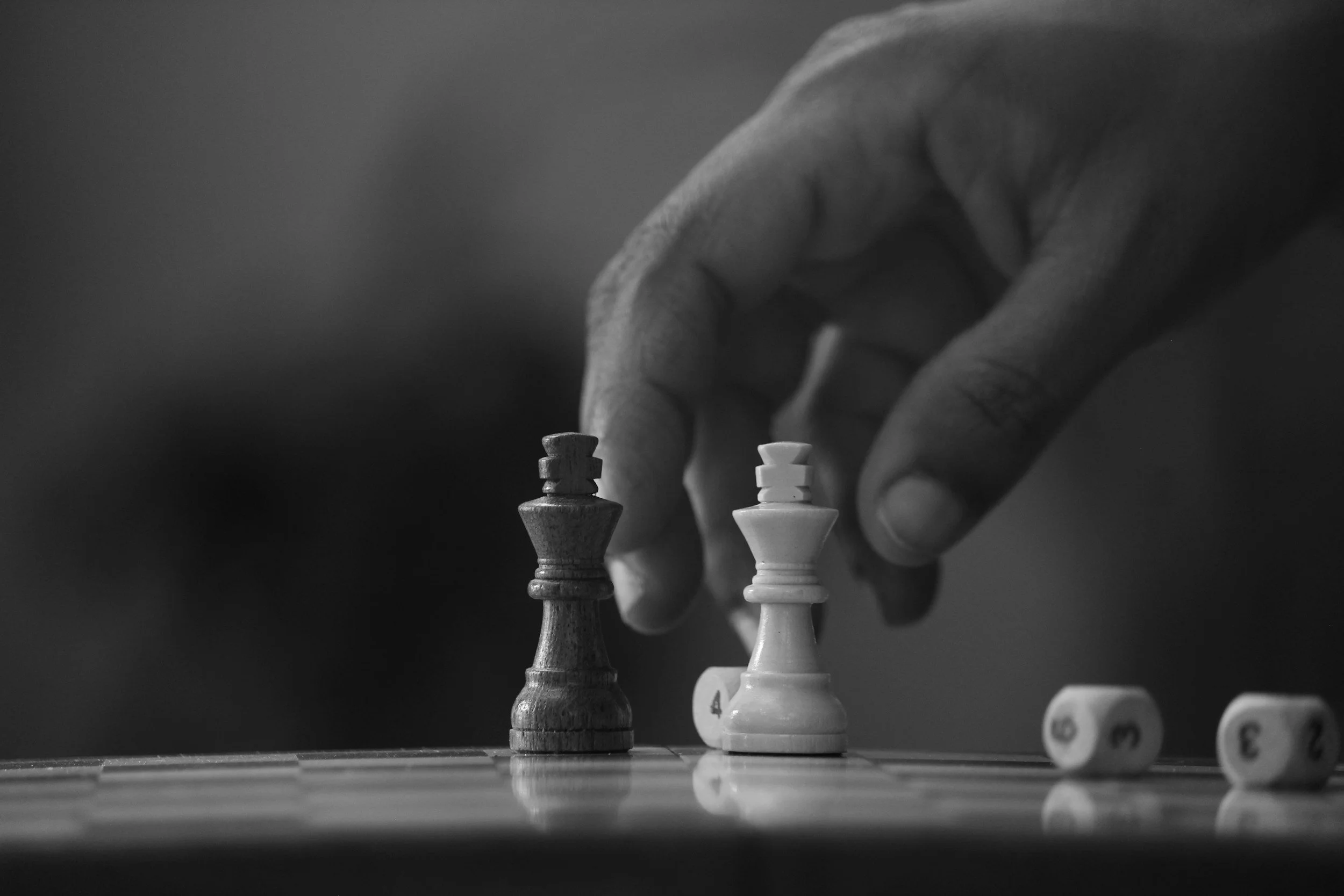 A black and white photo of a hand moving a chess piece on a chessboard, with two dice showing the numbers 3, 4, and 2.
