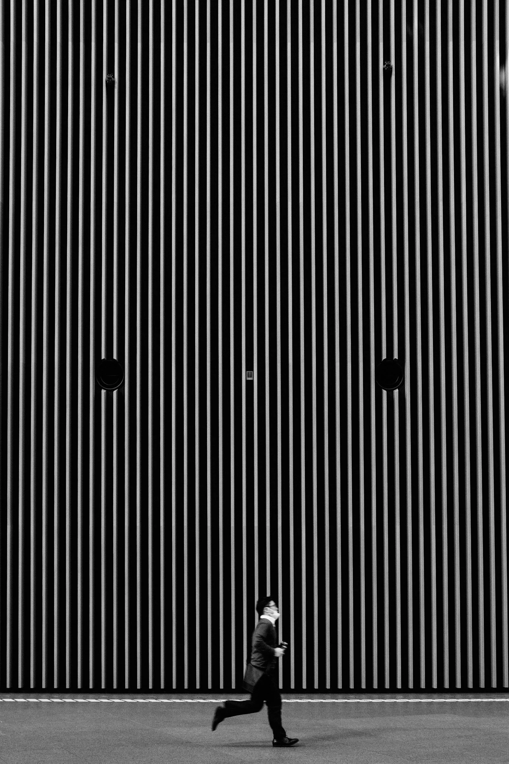 A person wearing a face mask, suit, and tie jogging past a large vertical striped wall in black and white.