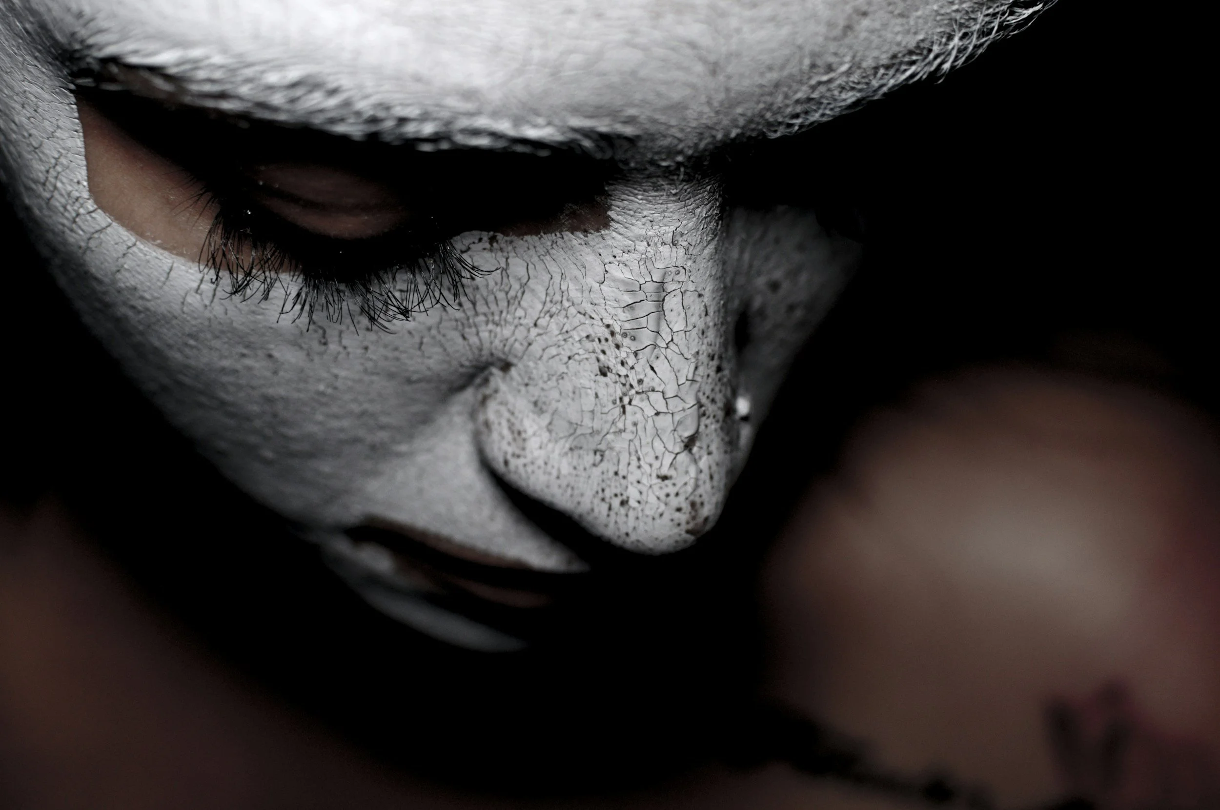 Close-up of a person's face with cracked white face paint, emphasizing the nose, eye, and eyelashes, with dark shadows highlighting facial contours.