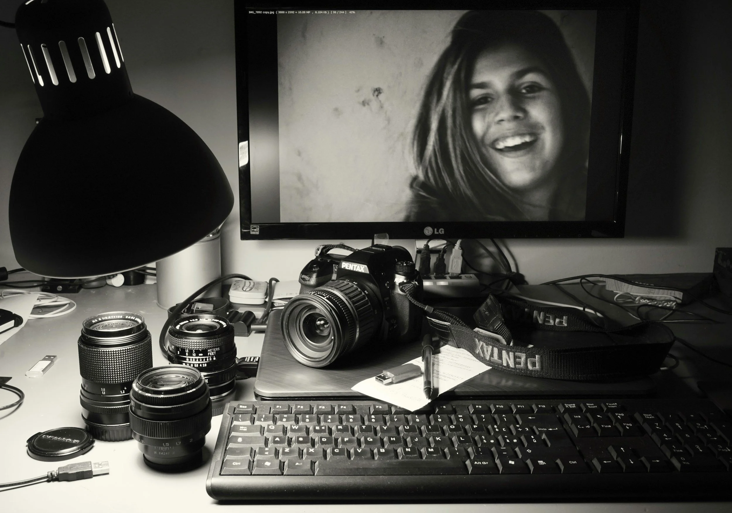 Photograph of a cluttered desk with camera gear, a monitor displaying a smiling woman, and a keyboard.