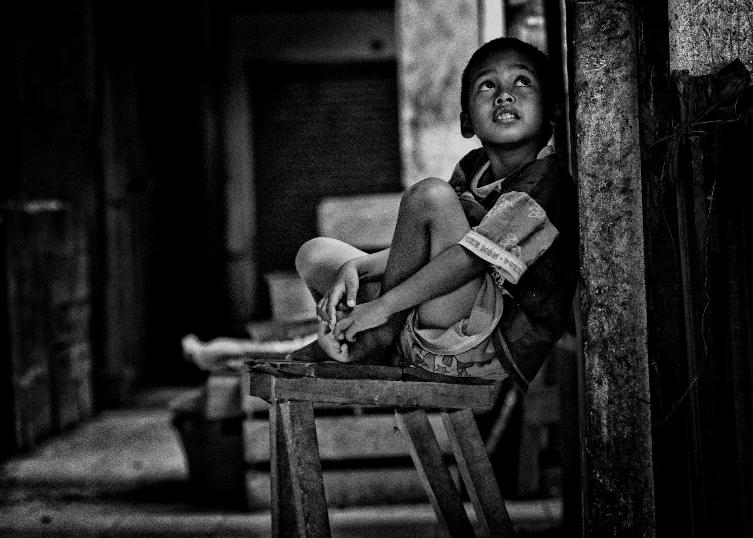 A young boy sitting on a small wooden stool, looking up with a thoughtful expression inside a rustic room.