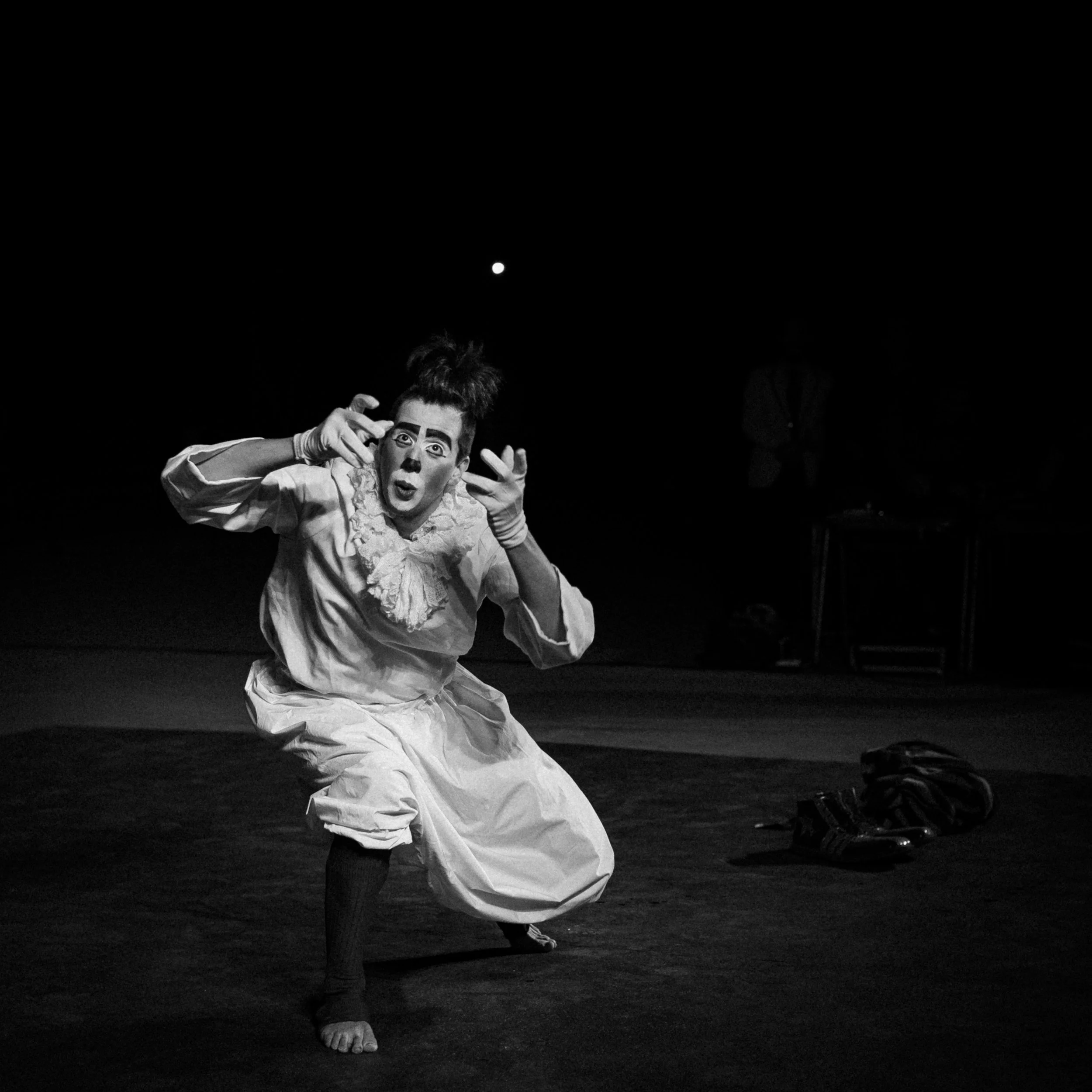 A mime performer dressed in vintage costume with exaggerated makeup, striking a dramatic pose on stage, with a dark background and a partially visible person in the distance.
