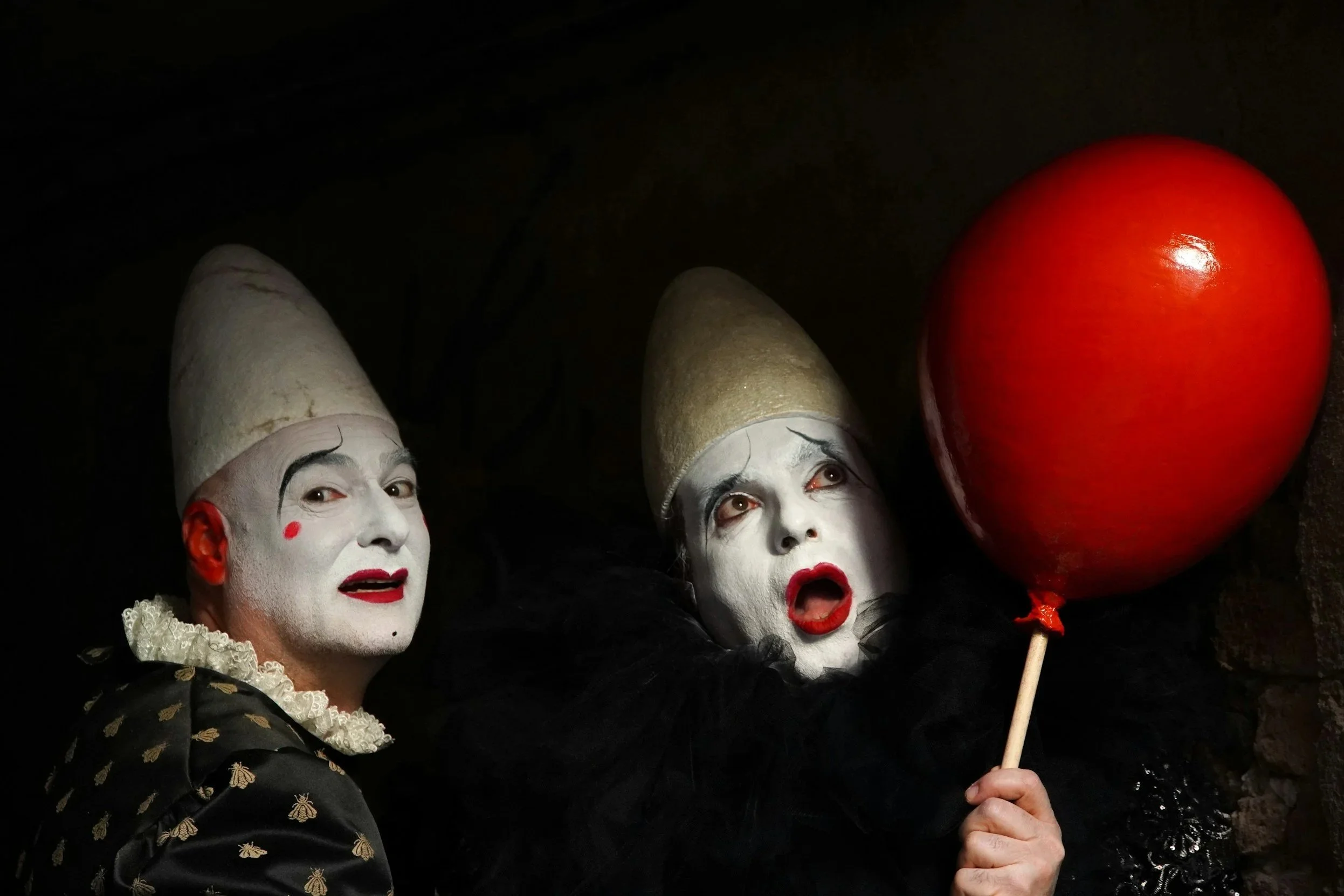 Two mime performers with white face paint, wearing pointed hats, dressed in black costumes, one holding a red balloon, set against a dark background.