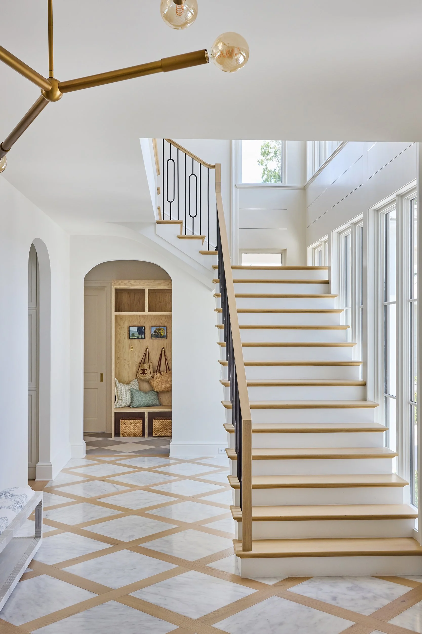 Interior of a modern home with a white staircase with wooden steps and a black metal railing, large windows, and a geometric patterned marble and wood floor.