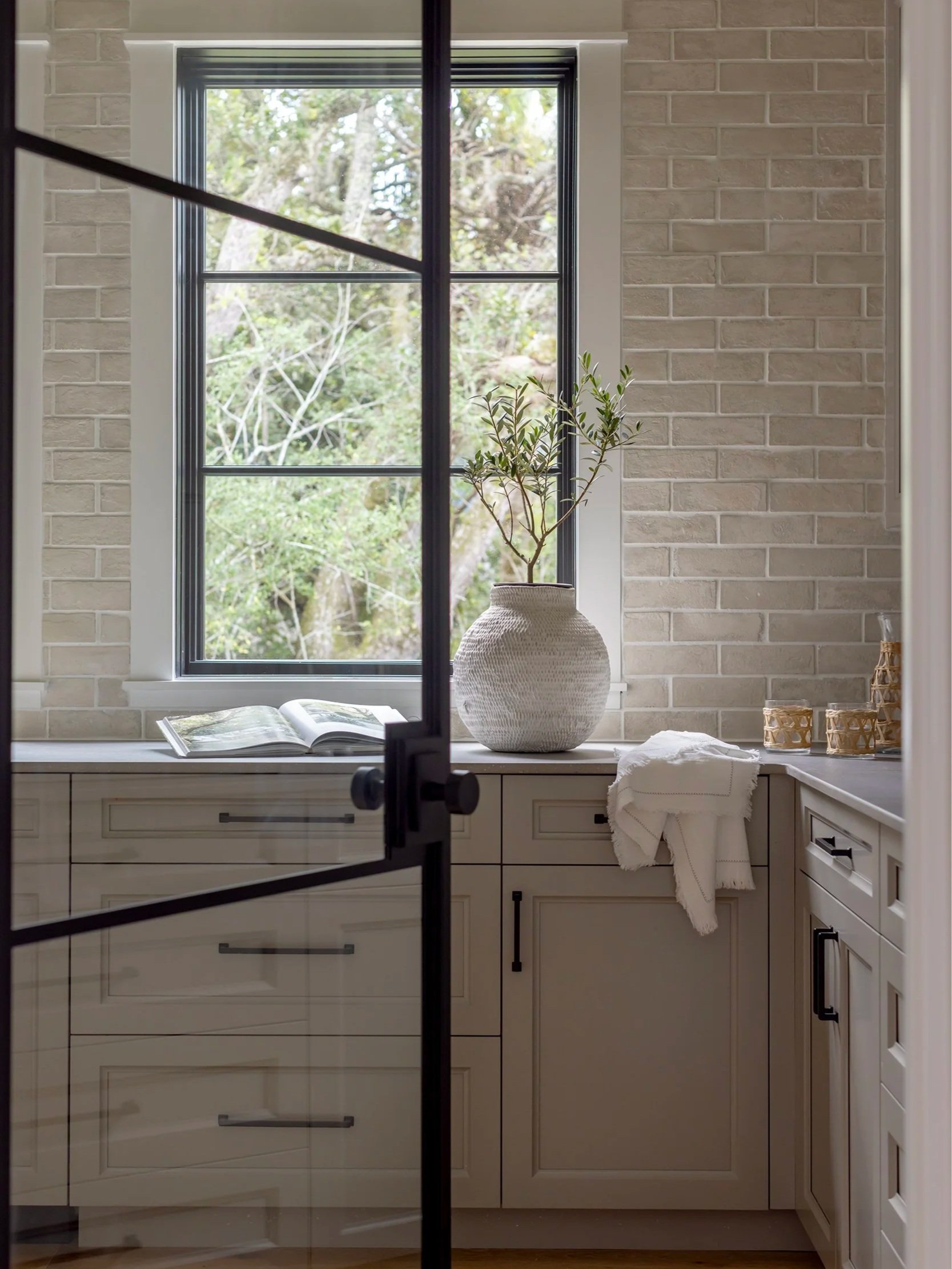 Kitchen with a large window, beige cabinets, open book, white ceramic vase with a plant, and wicker containers.