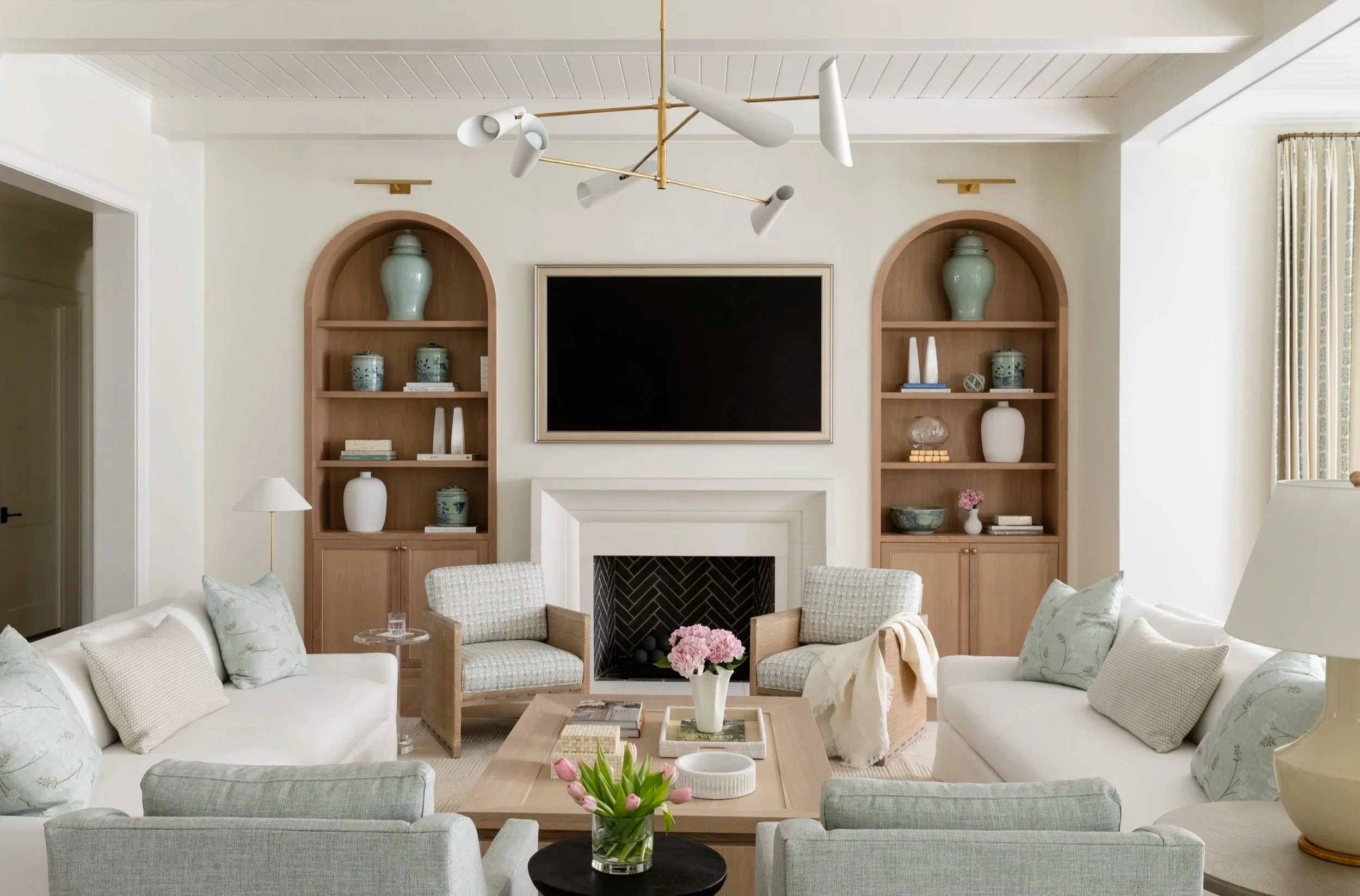 Living room with built-in wooden shelves, a large flat-screen TV above a white fireplace, sofas, armchairs, a coffee table with flowers, and a modern chandelier.