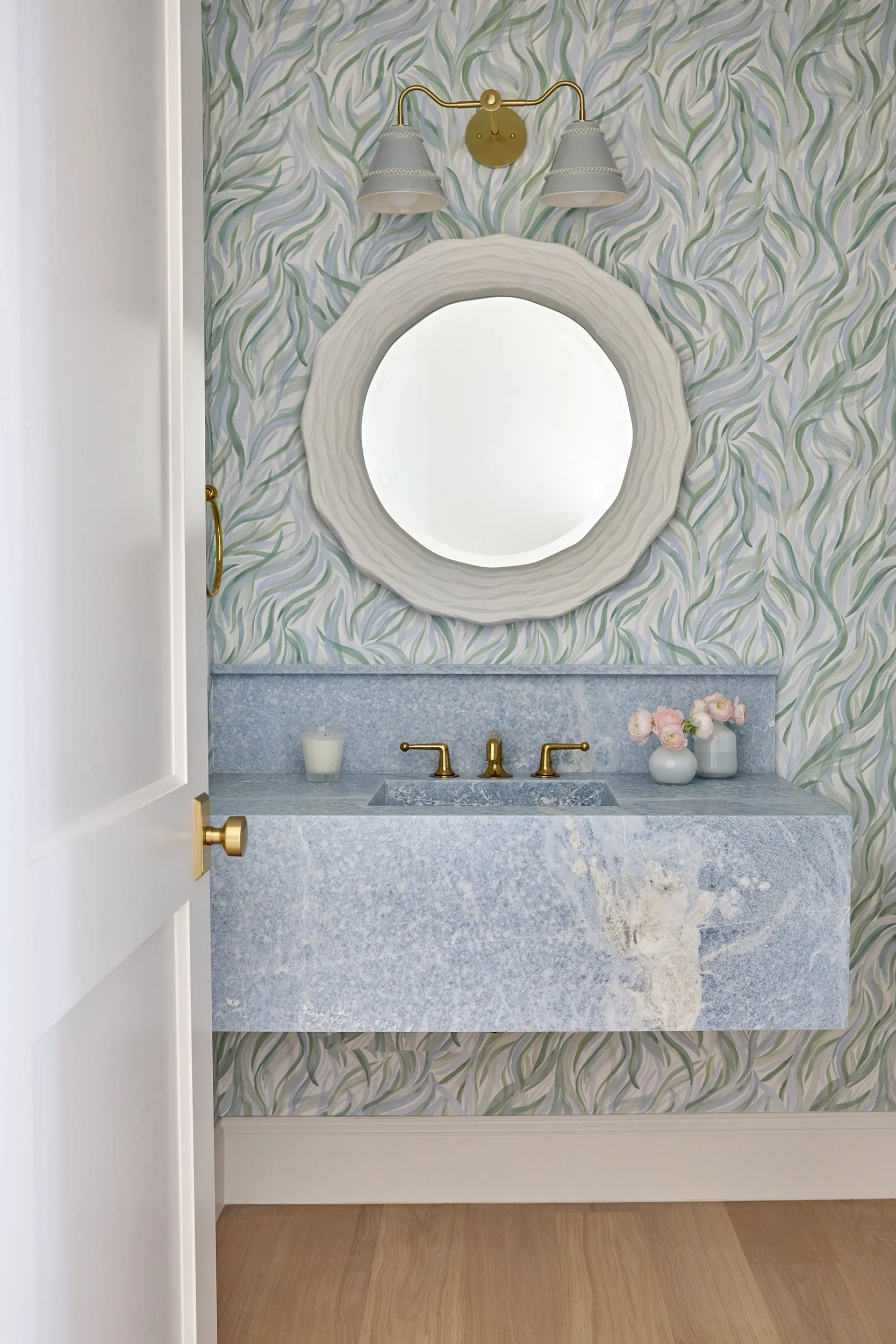A bathroom vanity with a blue-gray marble countertop, gold fixtures, a round mirror with a textured white frame, floral wallpaper, a small vase with pink flowers, and a candle.