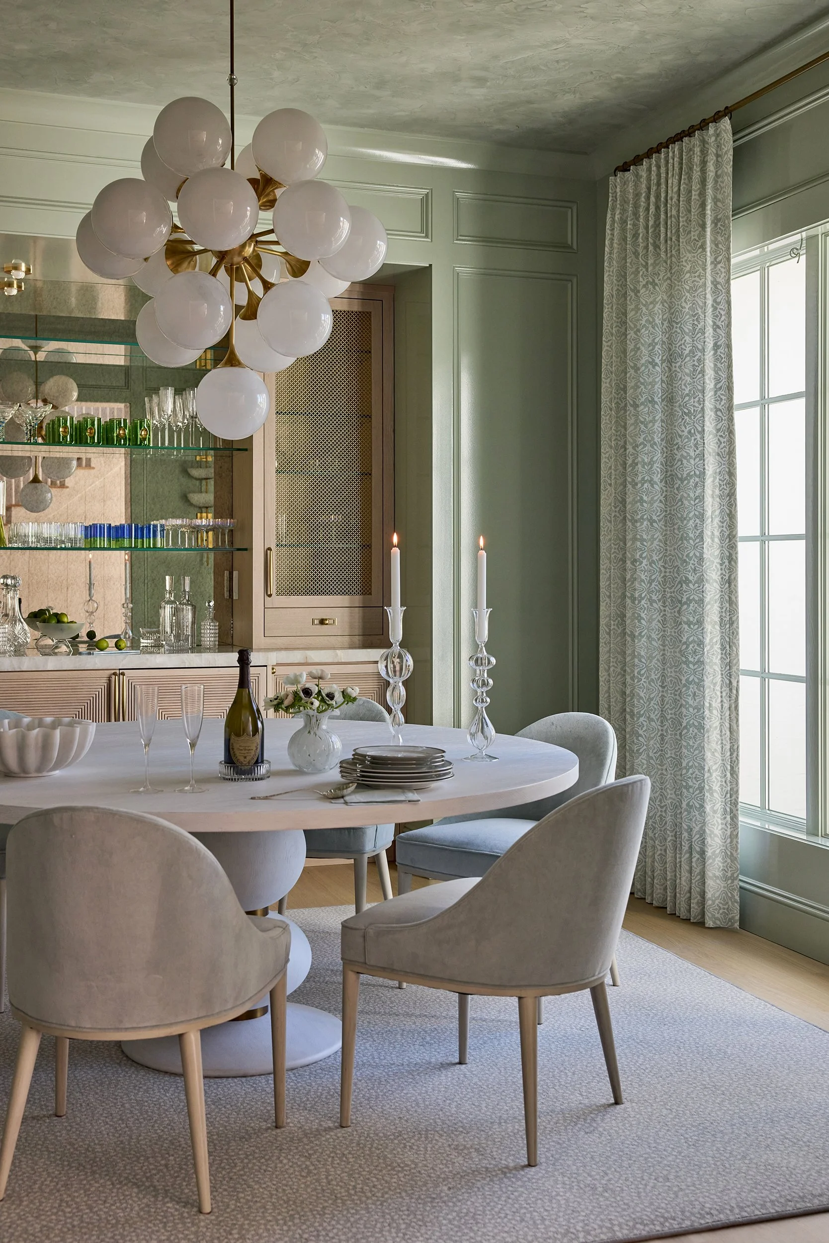 A dining room with a round table set with plates, glasses, a bottle of wine, and lit candles. There are six upholstered chairs around the table, a chandelier hanging above, and a large window with patterned curtains. A cabinet with glass shelves and 