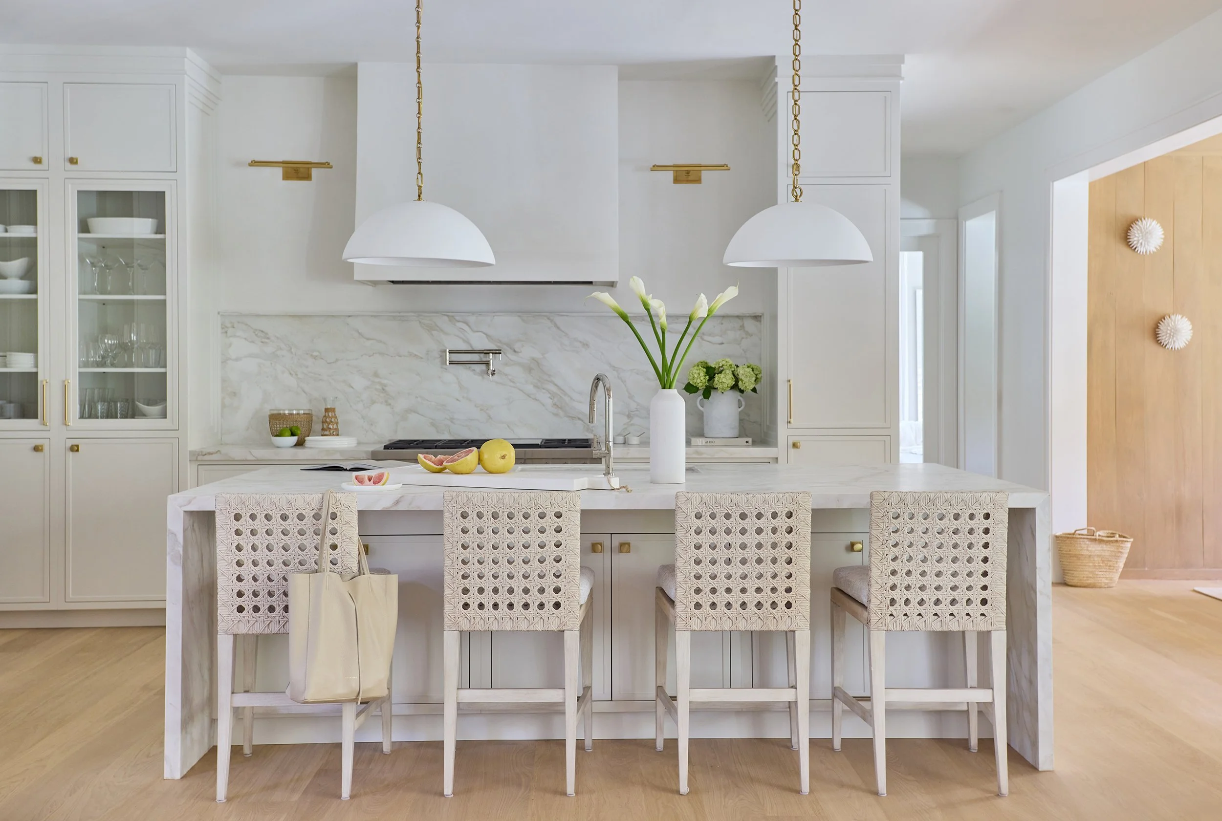 Bright white kitchen with marble island, four woven chairs, and gold accents. Potted flowers and fruits on the island.