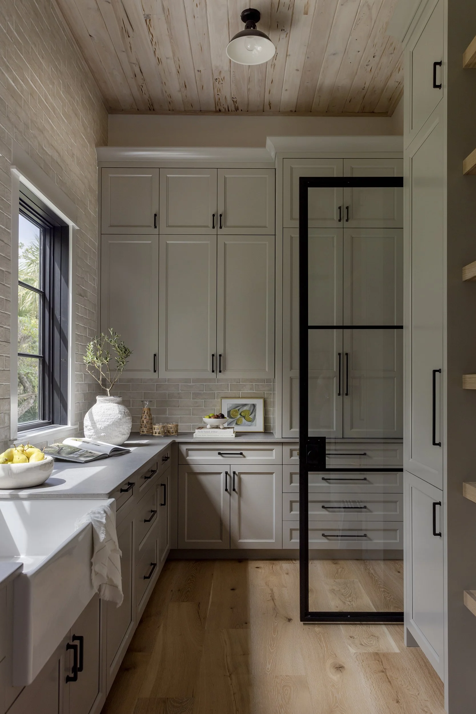 A modern kitchen with white cabinets, a black-framed glass door, wooden floors, a window with a view outside, and a wooden ceiling with a black and white ceiling light.