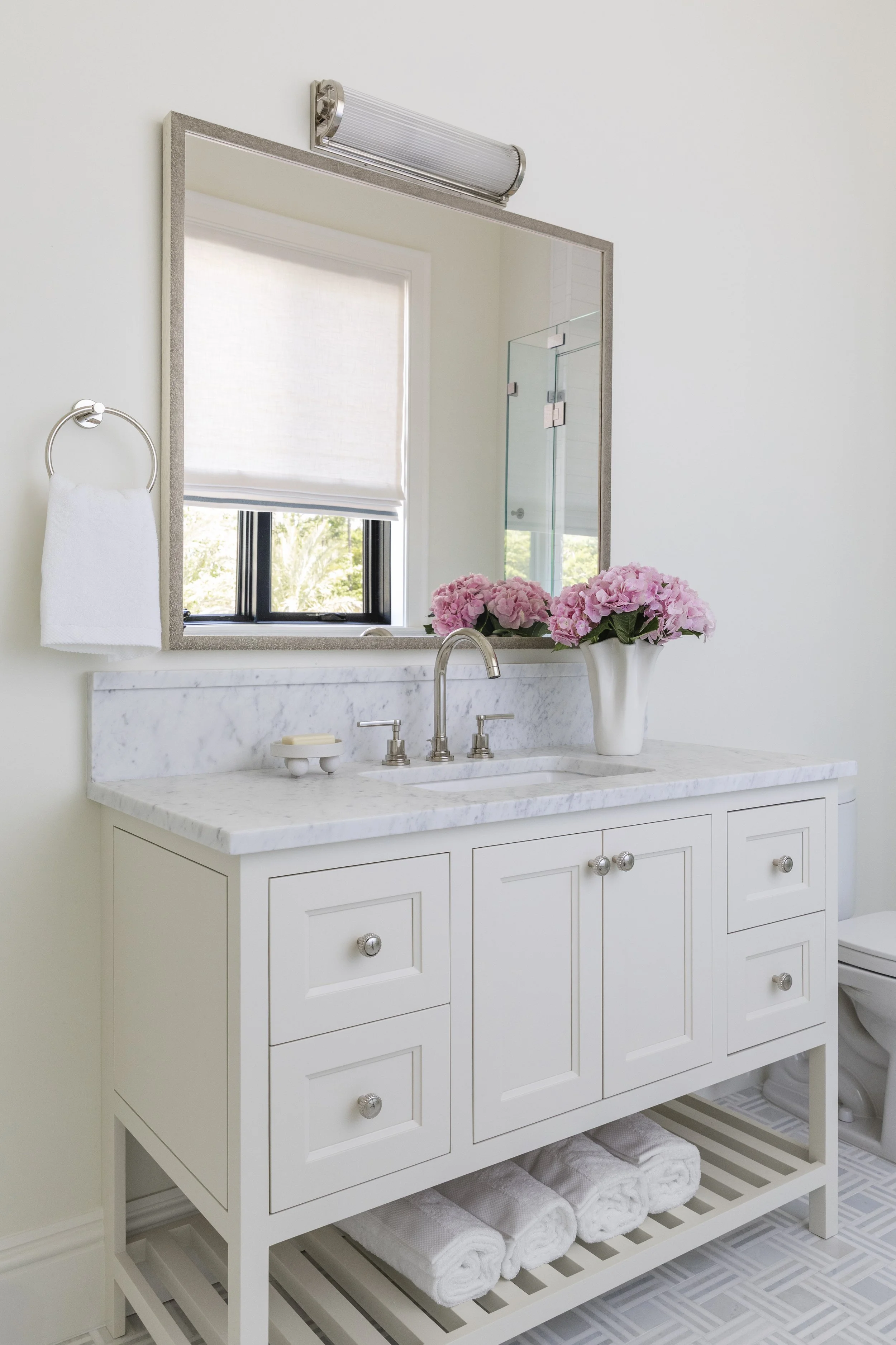 A white bathroom vanity with a marble countertop, a large mirror with a light fixture above, a vase of pink flowers, and a window with a roller shade. Towels are stored on the lower shelf.