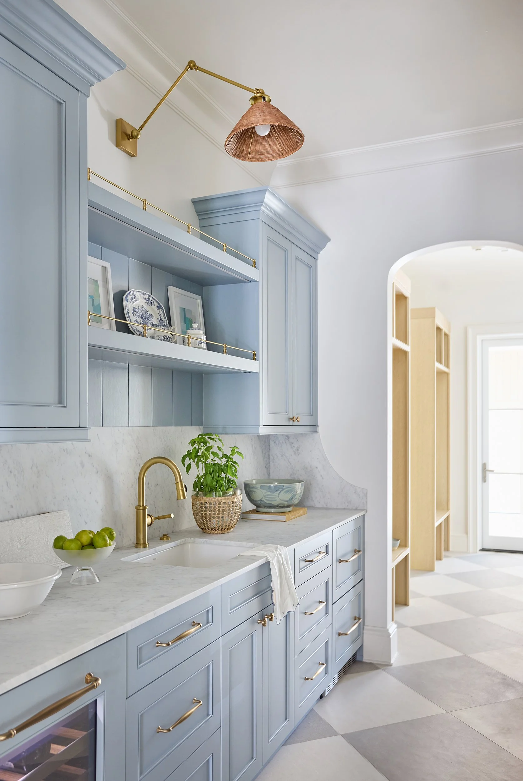 Light blue kitchen cabinets with brass handles, white marble countertop, open shelves with blue and white decor, a potted plant, a bowl of green apples, and a brass wall sconce with a rattan shade.