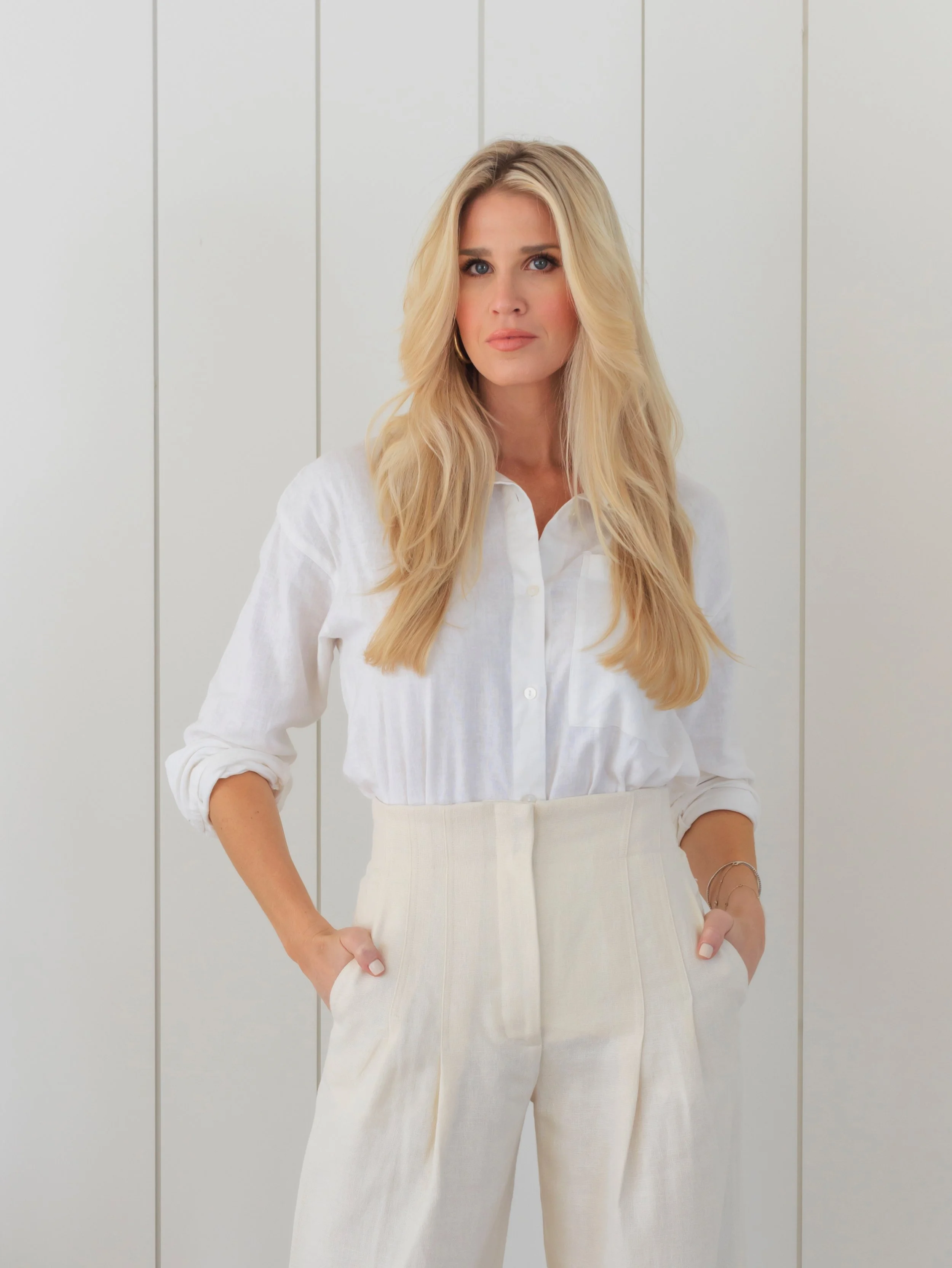A woman with long blonde hair wearing a white button-up shirt and beige high-waisted pants standing against a plain background.