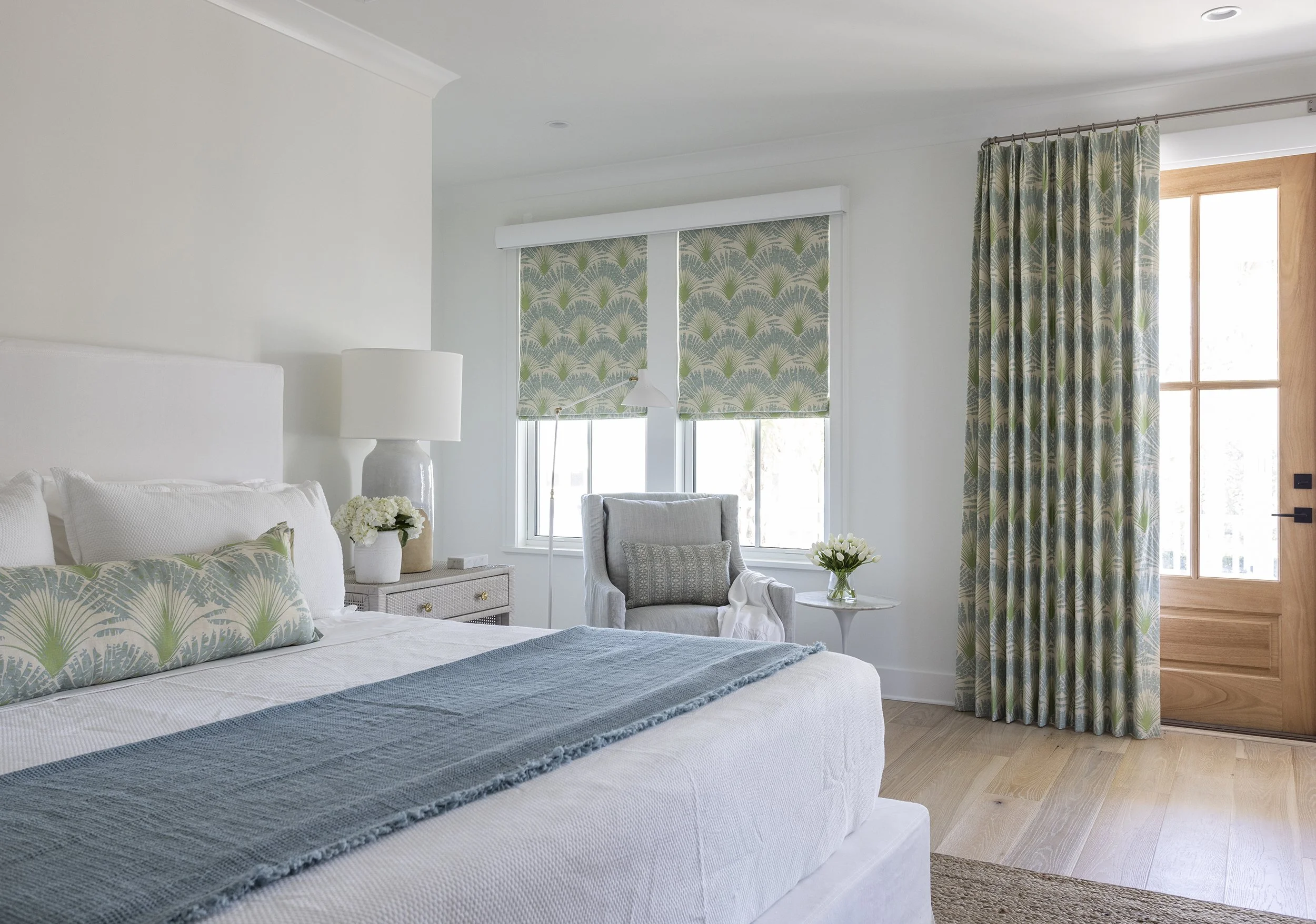 A bedroom with white walls, a large bed with white bedding and decorative pillows, a nightstand with a lamp and flowers, a gray armchair with cushions, a window with patterned blinds, and a door with patterned curtains.