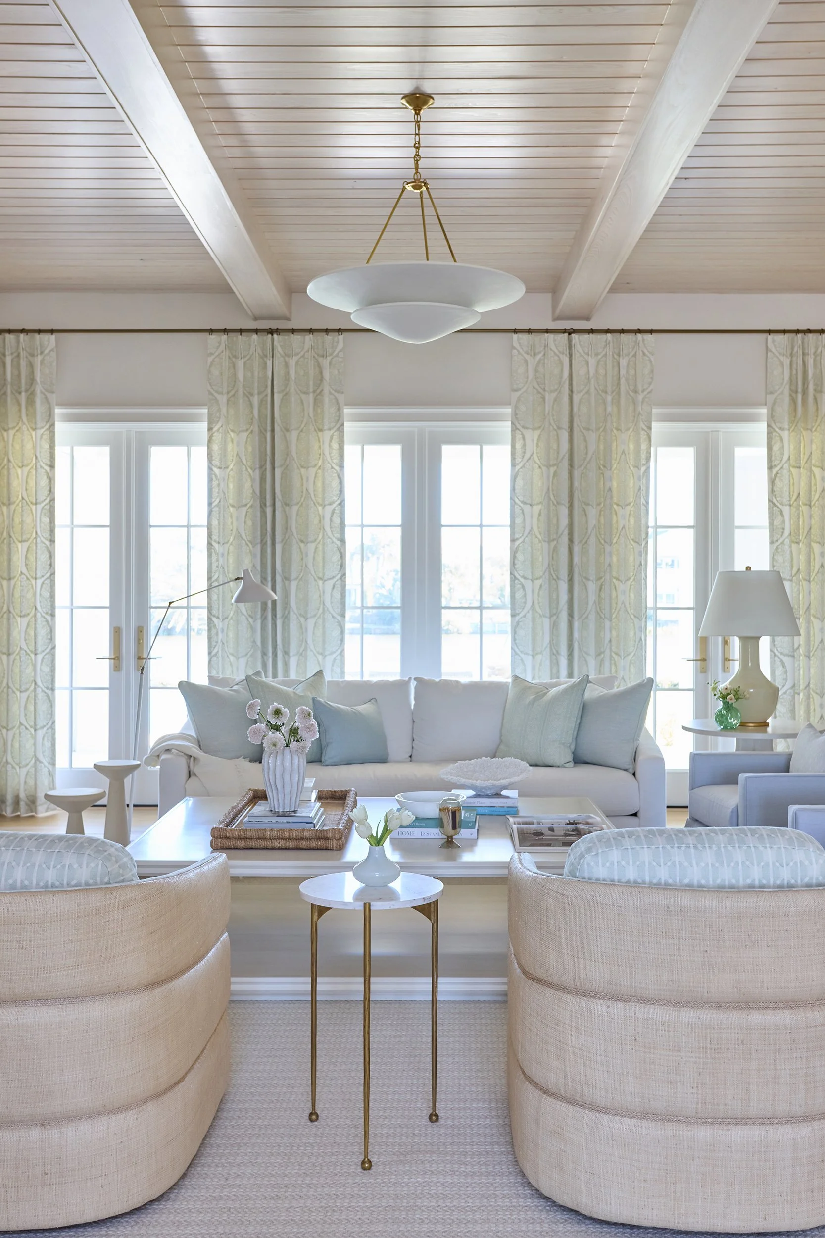 Bright living room with beige upholstered chairs, white sofa, large windows with patterned curtains, and light-colored decor.