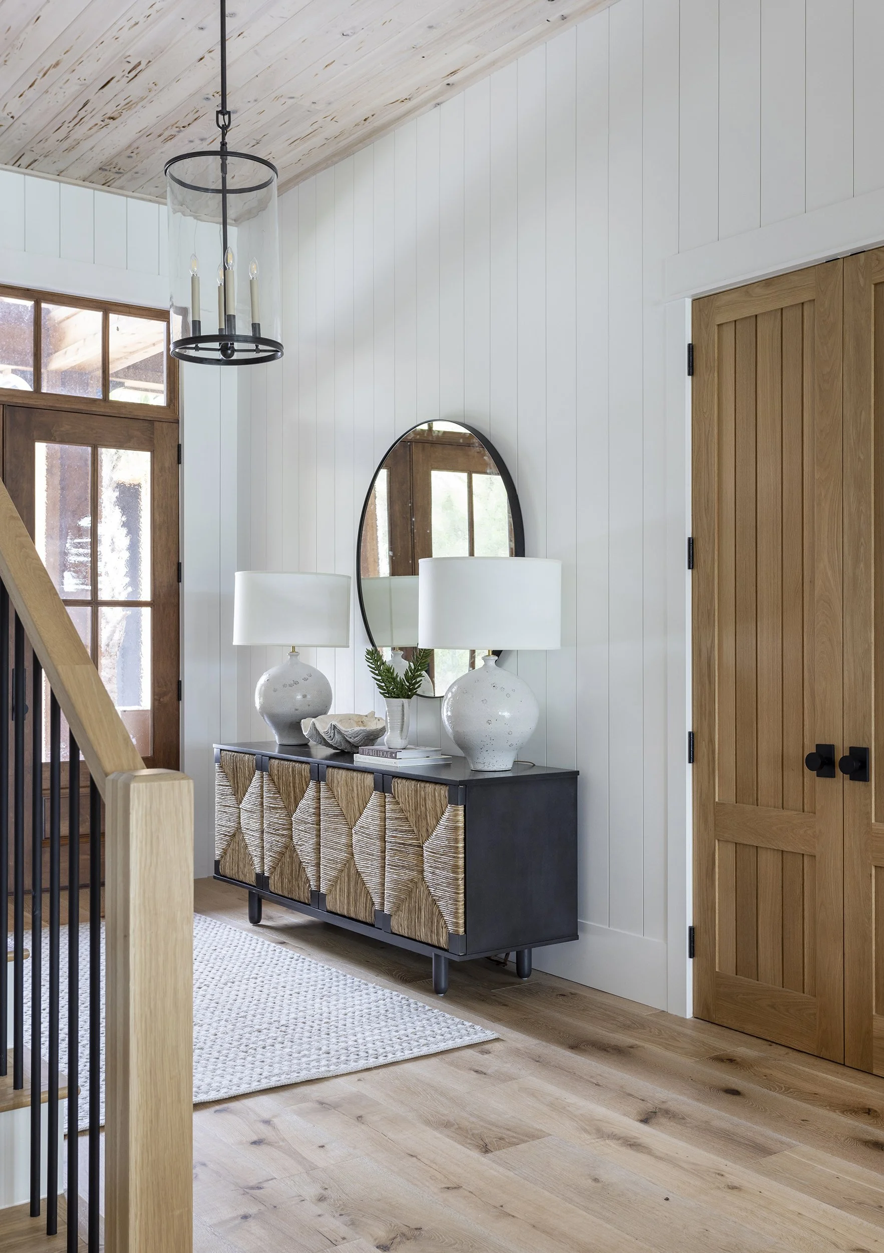 Entryway with a black sideboard, wicker-front cabinet, two white table lamps, and a mirror on a white beadboard wall, with wooden doors and a staircase.