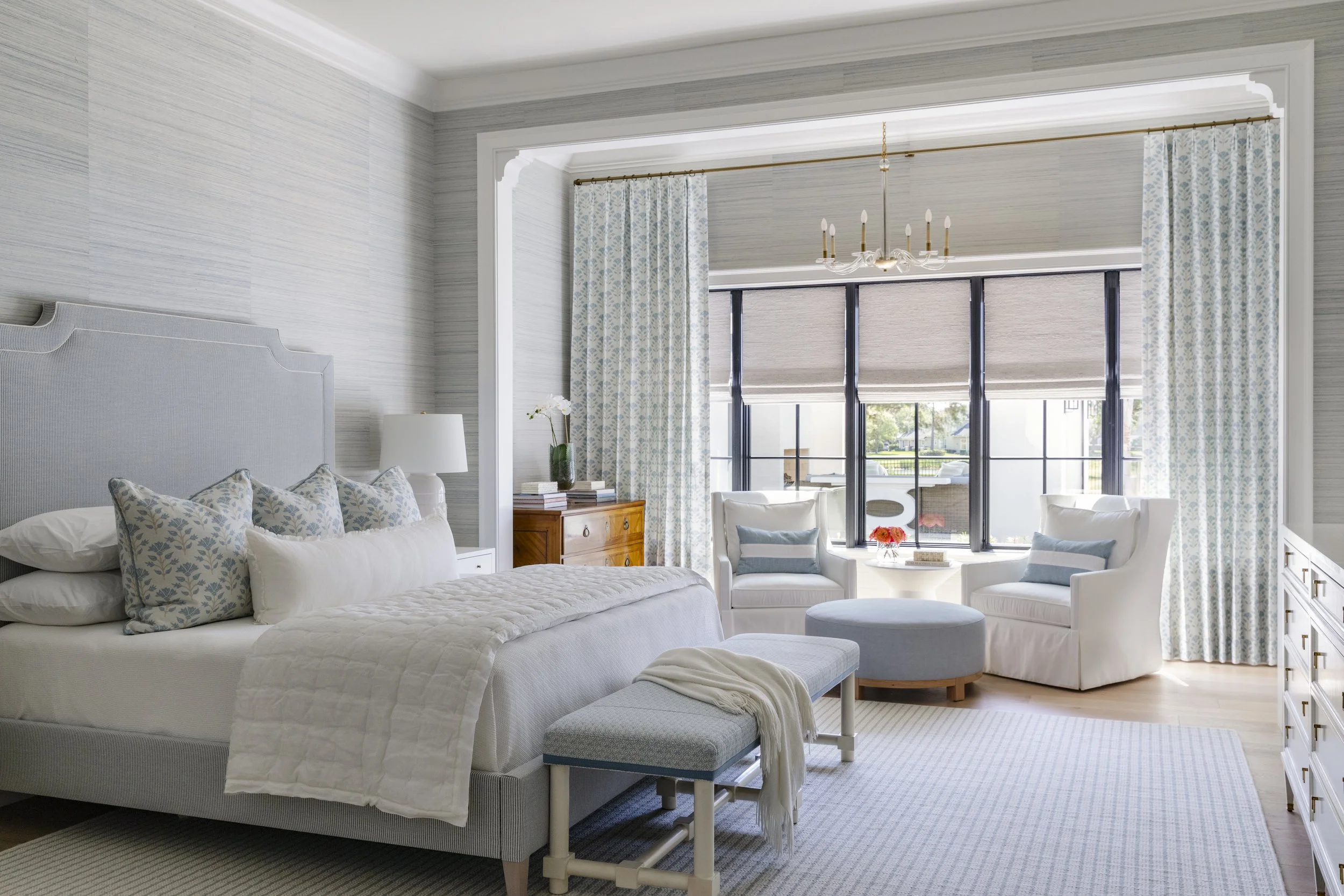 Bright bedroom with light gray walls, a large bed with a light gray headboard, white bedding, and multiple decorative pillows with light blue and beige patterns. There is a white bench at the foot of the bed with a white throw. To the right, a wooden