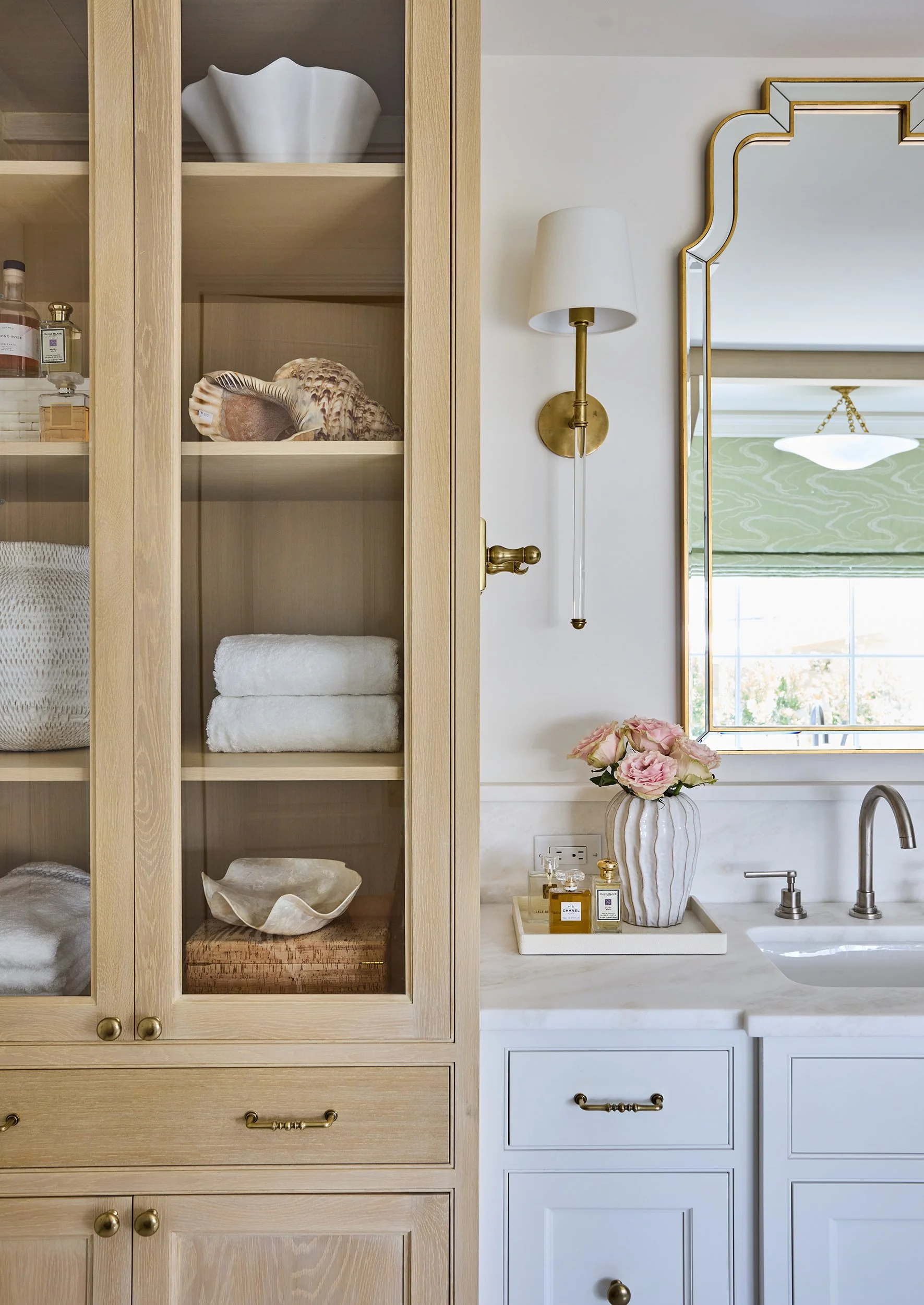 A bathroom with a light wood cabinet on the left containing towels and decorative items. To the right, a white vanity with a marble countertop, a sink, and a mirror. Above the vanity, a brass wall sconce and next to it, a mirrored wall with gold trim