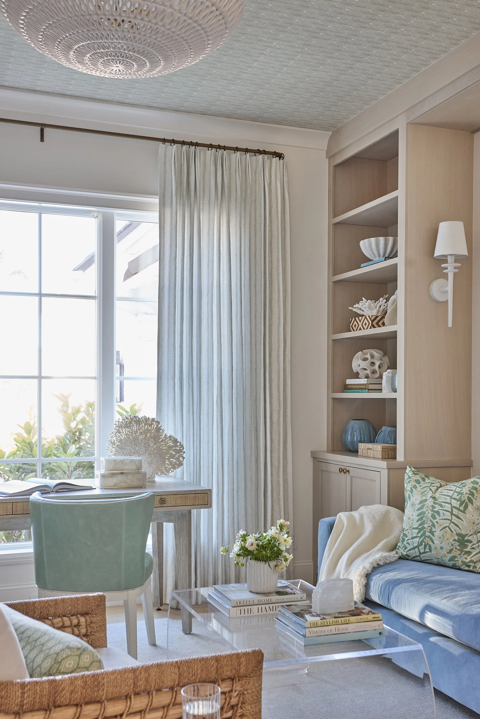 Cozy living room with a window, curtains, and decorative shelves with ceramics and books, a coffee table with flowers, and a sofa with cushions.