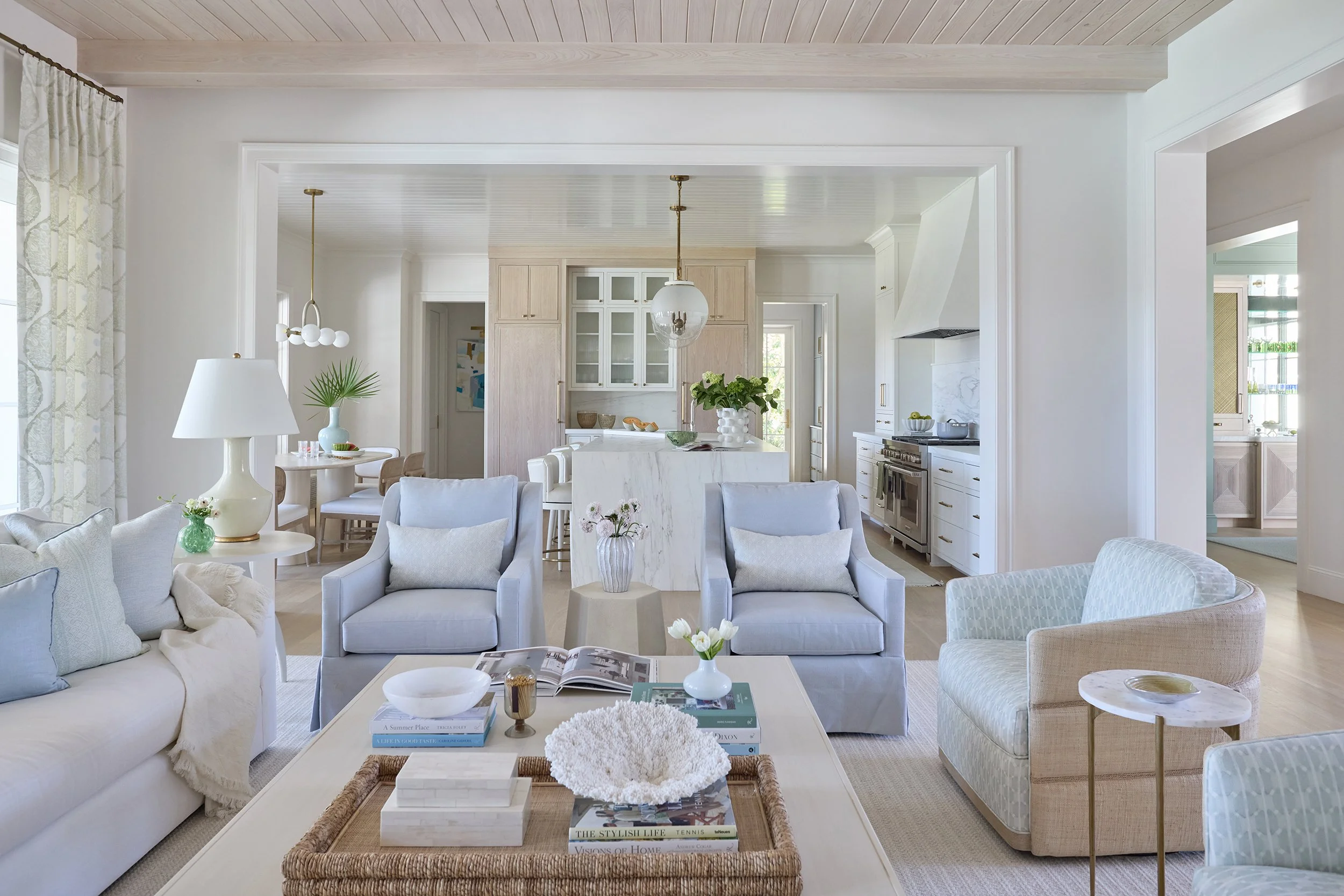 Bright, airy living room and open kitchen with white and neutral-toned furniture, a central island, and decorative plants.
