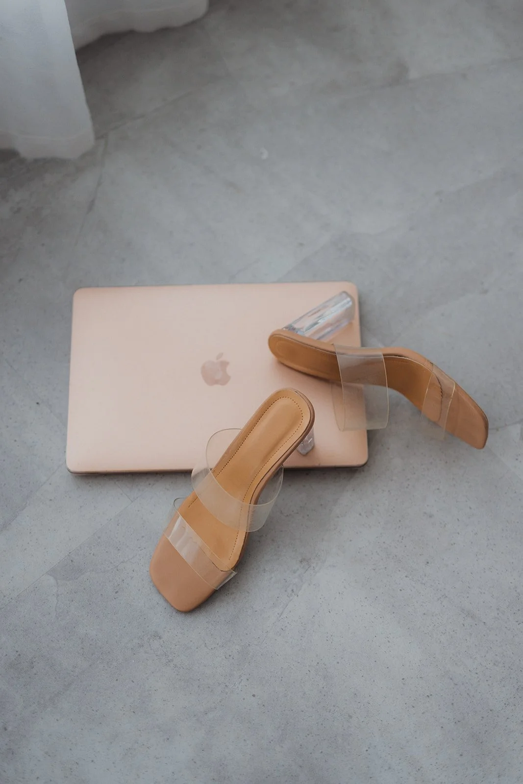 Ein aufgeschlagener, rosa MacBook mit einem Paar beiger, transparenter Seek-Absatzsandalen darauf, auf einem grauen Boden.