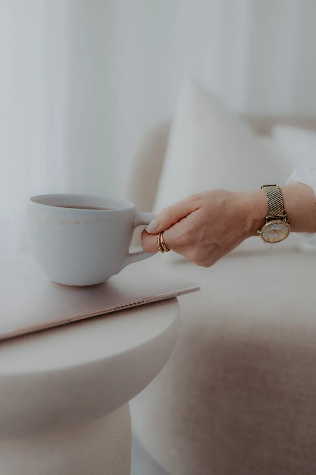 Eine Hand hält eine weiße Kaffeetasse mit Kaffee, auf einem Tisch. Die Person trägt eine goldene Uhr und mehrere Ringe.