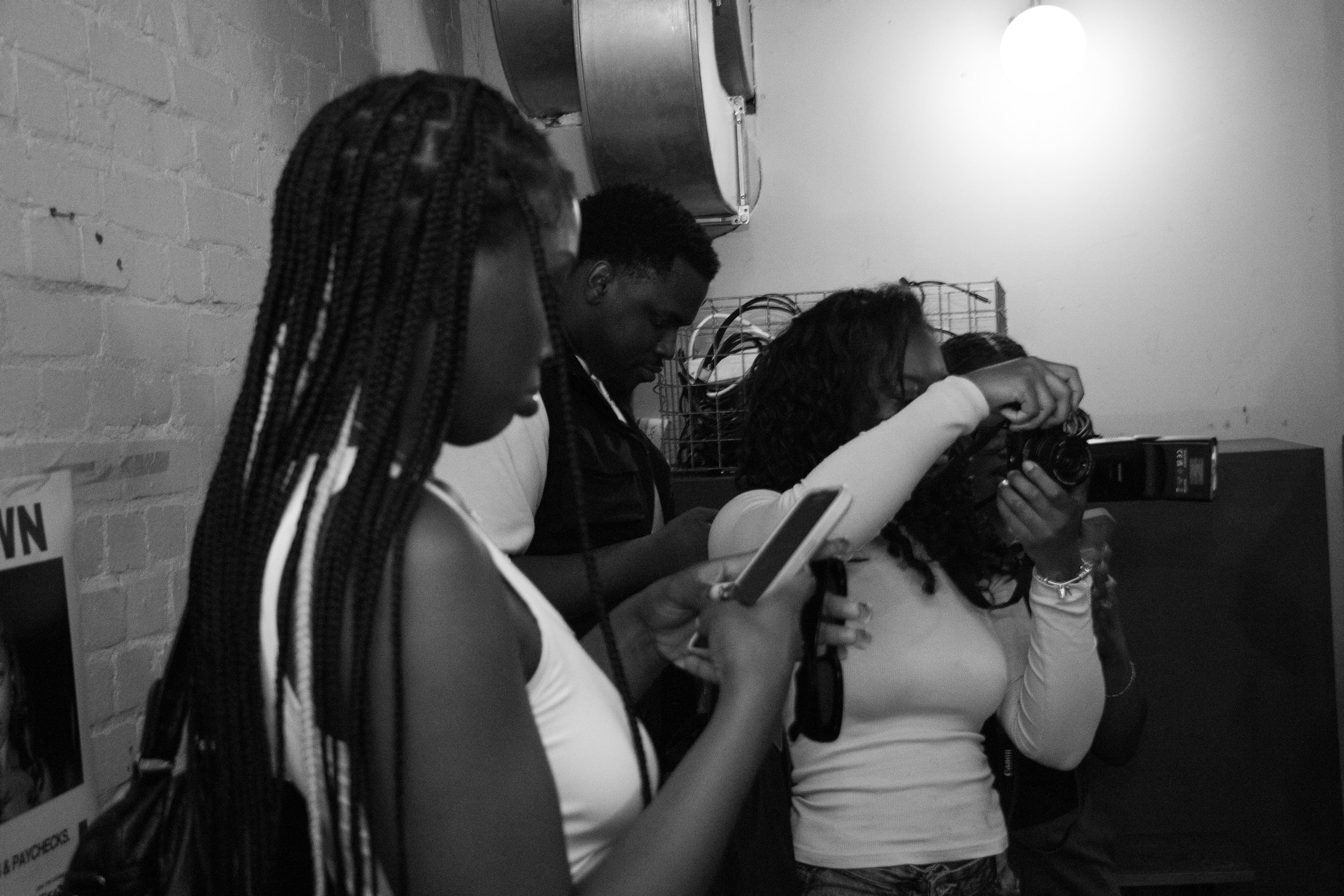 Four people standing close together, looking at their phones or camera, indoors with a brick wall and a light on the ceiling.