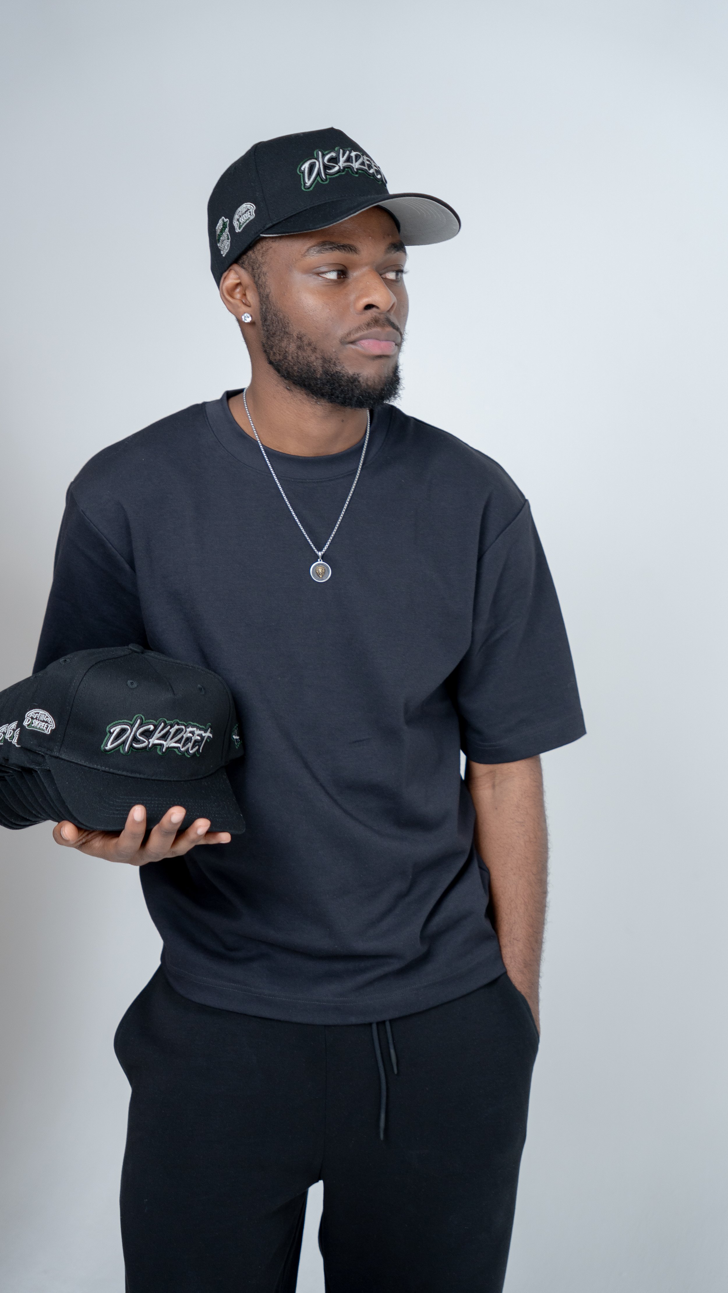 A young man dressed in black, wearing a black cap and holding a black baseball cap with the logo 'Diskreet', standing against a plain white wall.