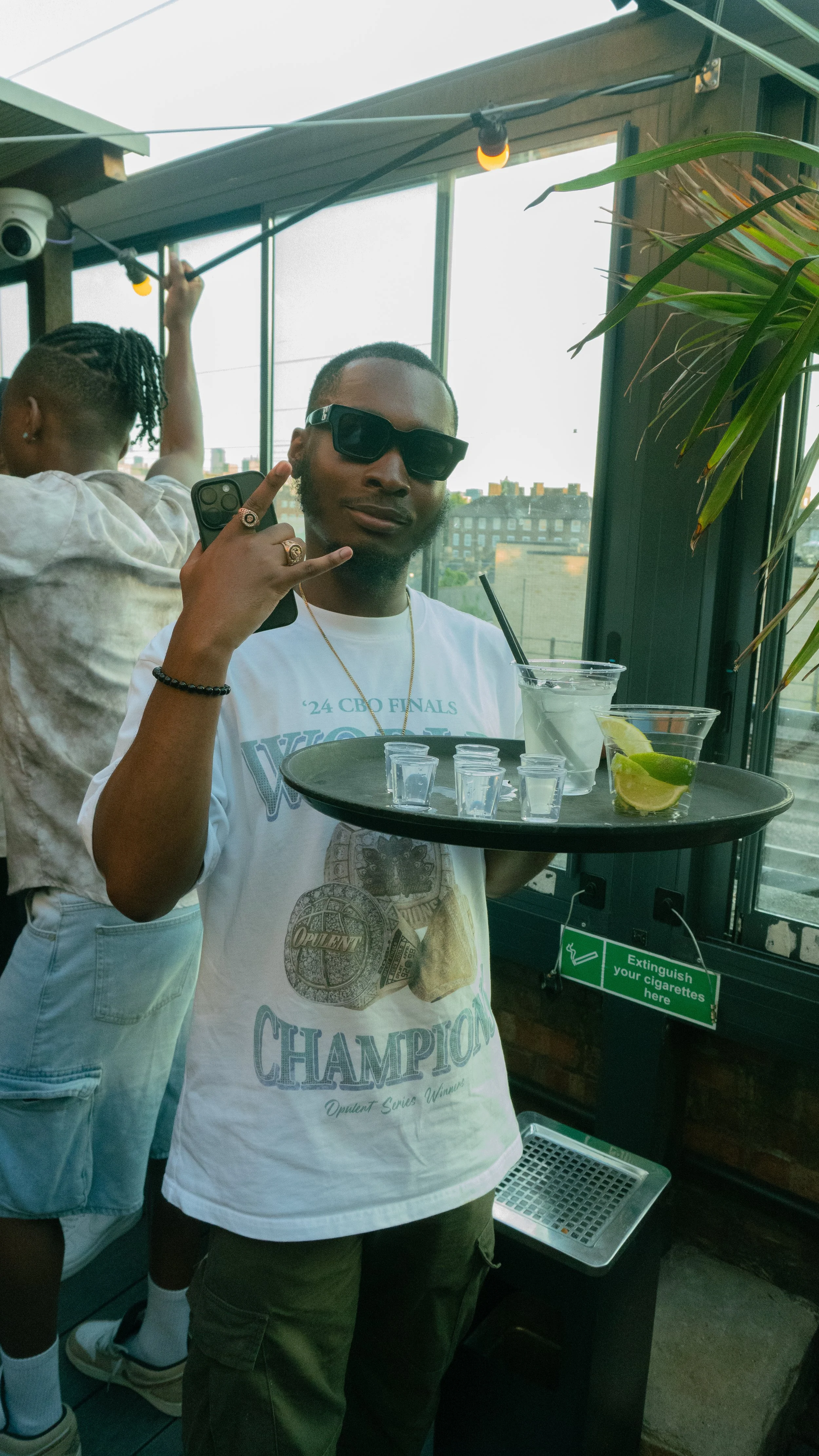 Man wearing sunglasses holding a tray with drinks, lemons, and small shot glasses inside a restaurant or bar.
