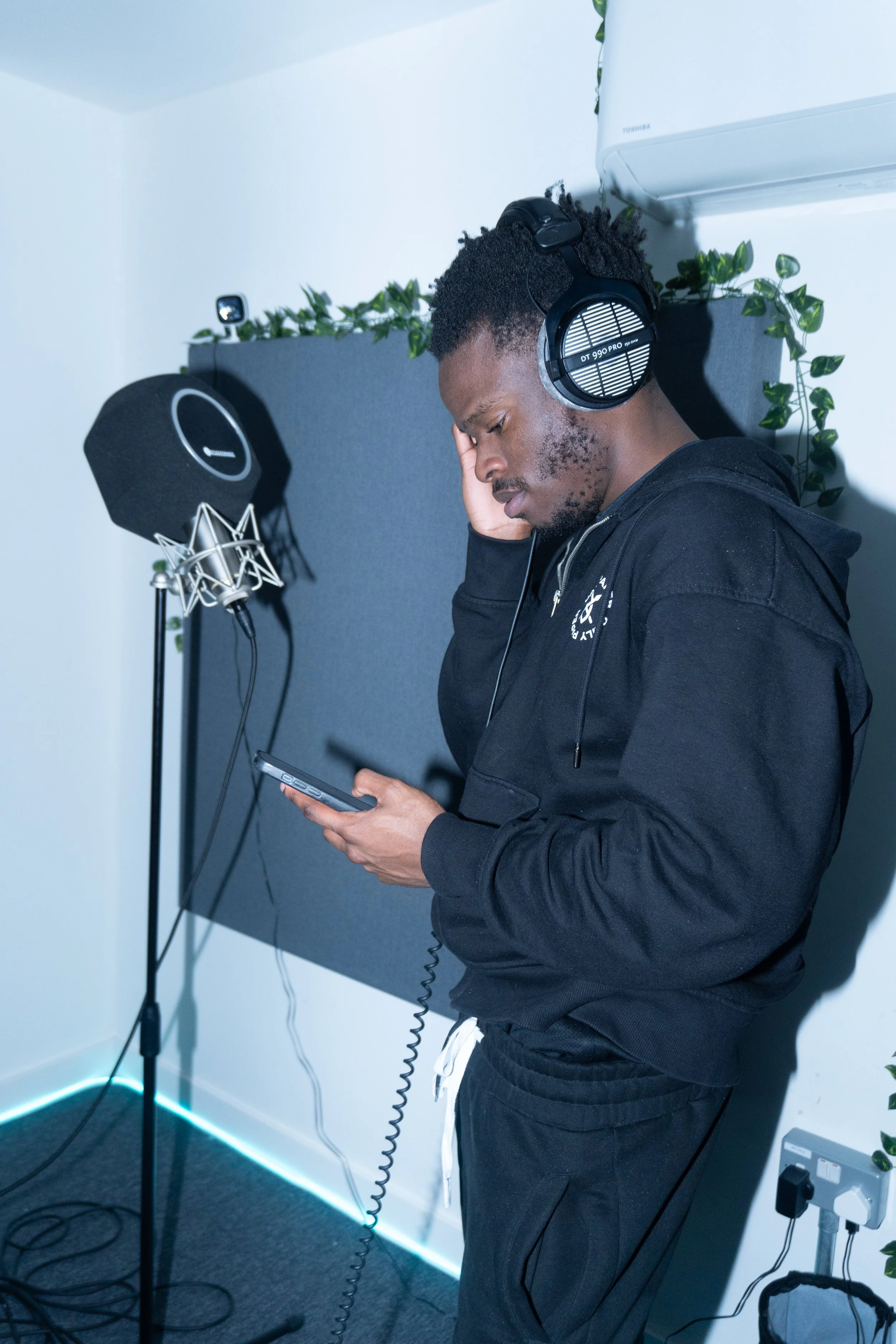 A man in a black hoodie and black sweatpants wearing large headphones stands in a recording studio, holding a phone and looking down at it.
