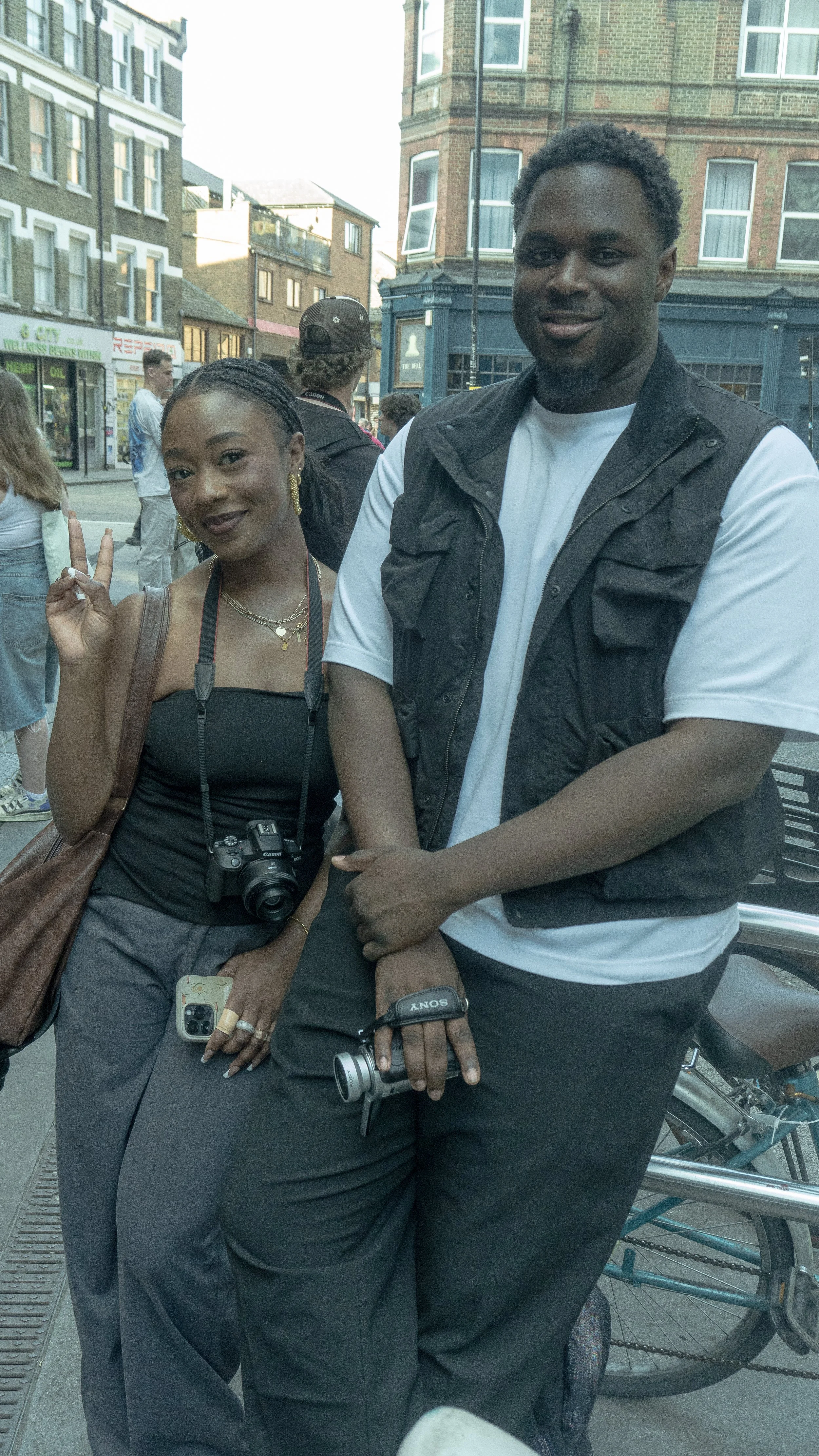 Two people standing close together on a city street, the woman on the left making a peace sign, wearing a black top, gray pants, and jewelry, with a camera around her neck and a phone in her hand. The man on the right is wearing a white shirt and black vest, with his arms crossed. Background includes other pedestrians and brick buildings.