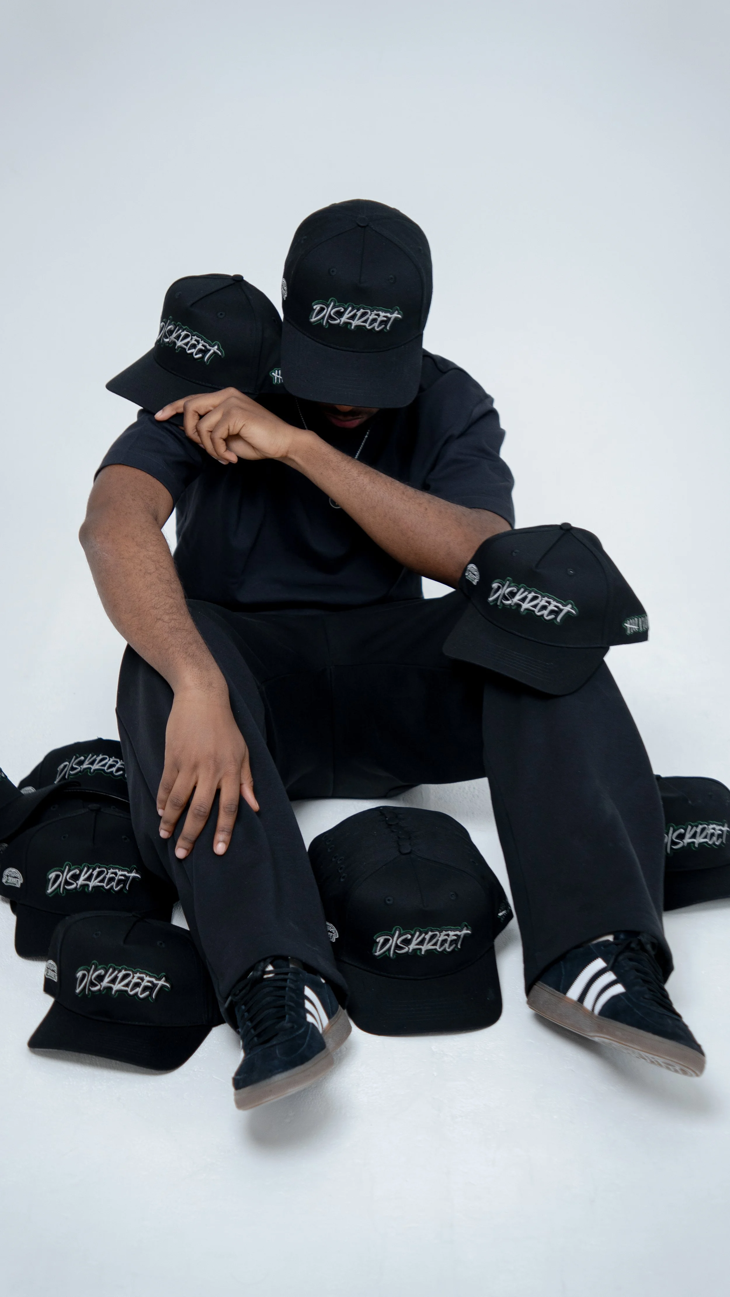 A person wearing a black cap, shirt, and pants, surrounded by multiple black caps, all with a logo that says 'DISKREET'. The person is sitting on the floor with one hand resting on their knee and the other adjusting the cap on their head, in a studio with a plain white background.