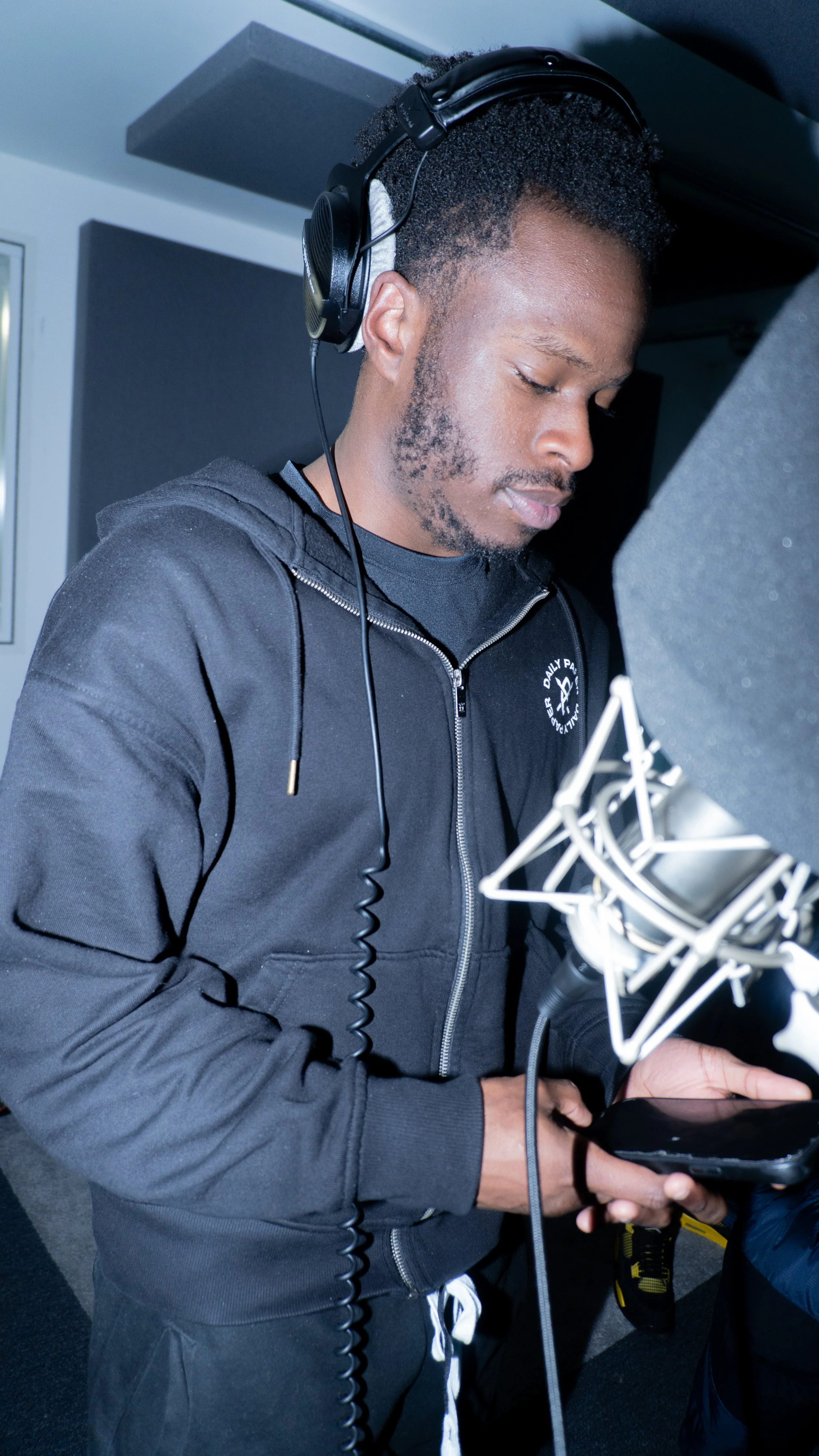 A man in a black hoodie with headphones in a recording studio, speaking into a microphone while looking at a phone.