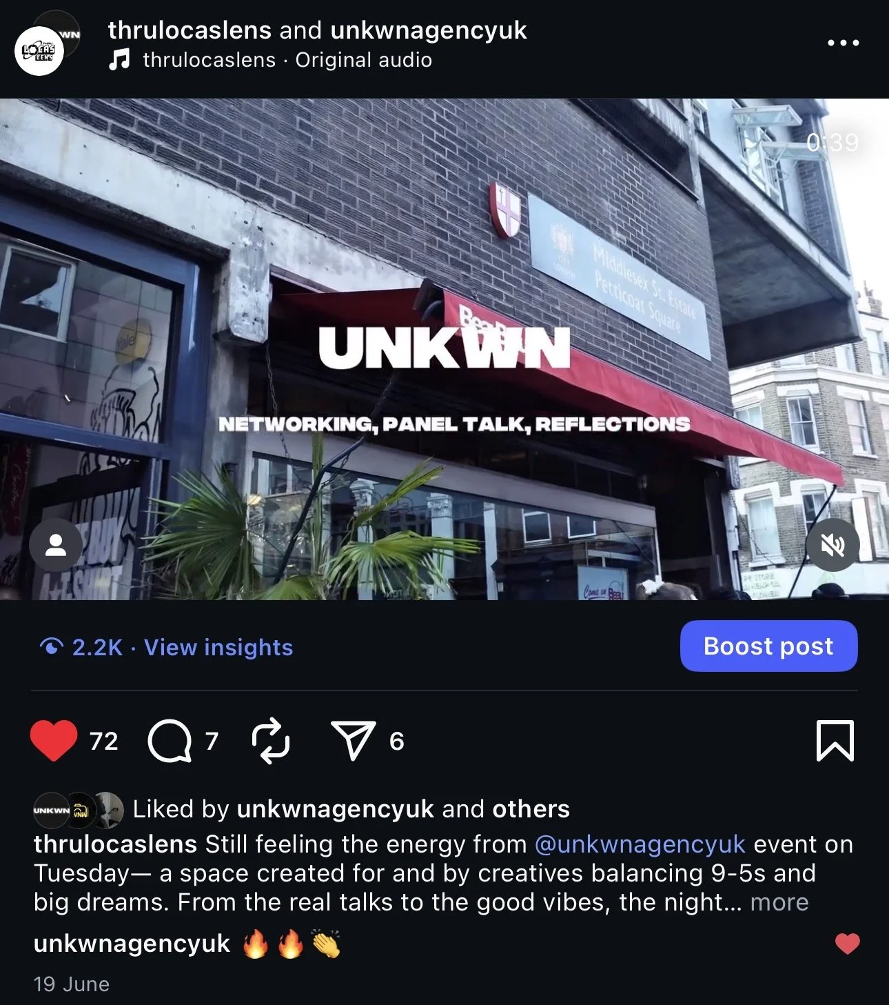 Exterior of a building with a red awning that has the UNKWN logo on it. A couple of small signs can be seen on the building's wall, including a blue street sign with white text. There are some plants in the foreground and other buildings nearby.