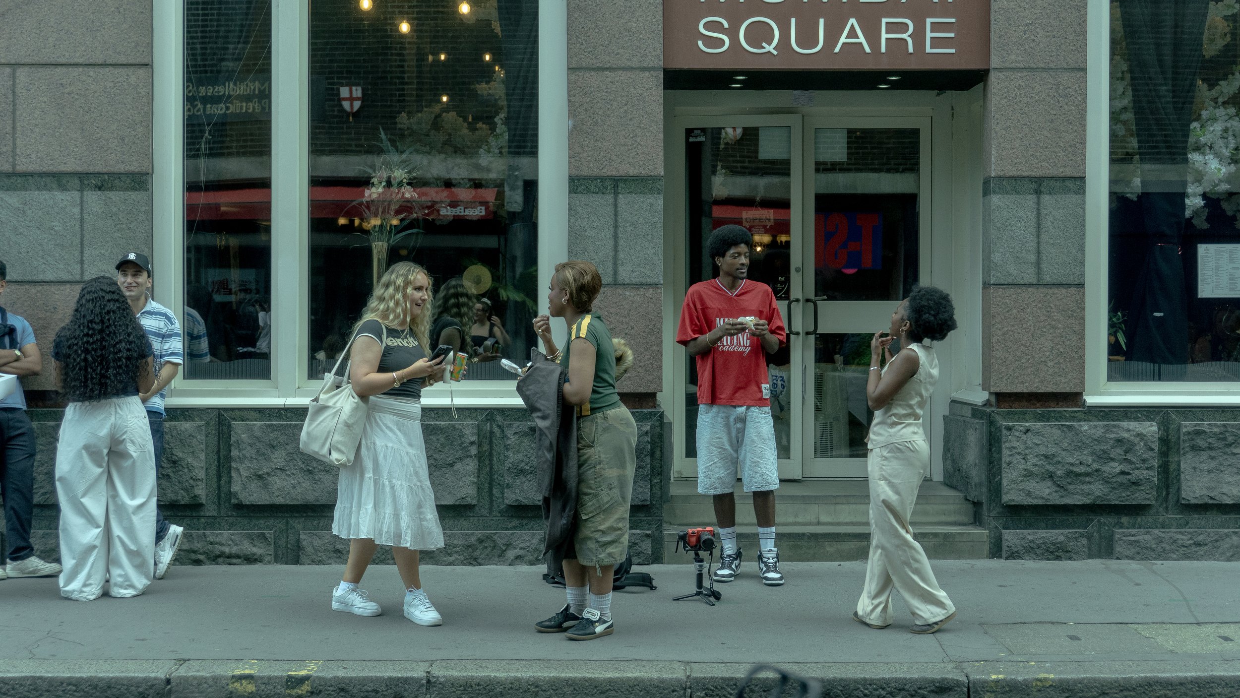 A group of people standing and chatting outside a building with a sign reading 'SQUARE'. Some people are taking photos or holding objects, and one person has a camera on the ground.