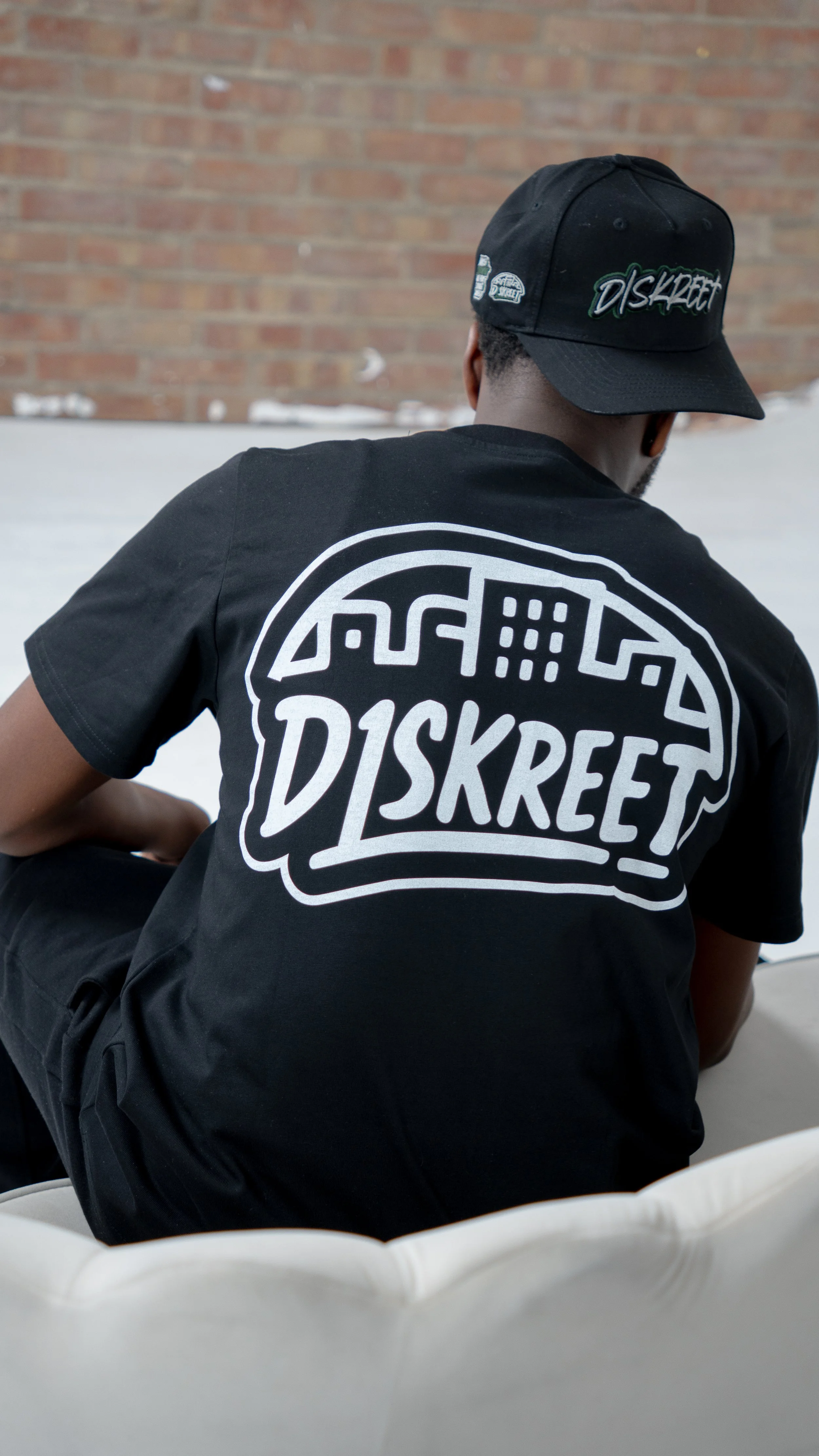 Back of a man wearing a black T-shirt and black cap, both with the 'DISKREET' logo, sitting on a white couch against a brick wall.