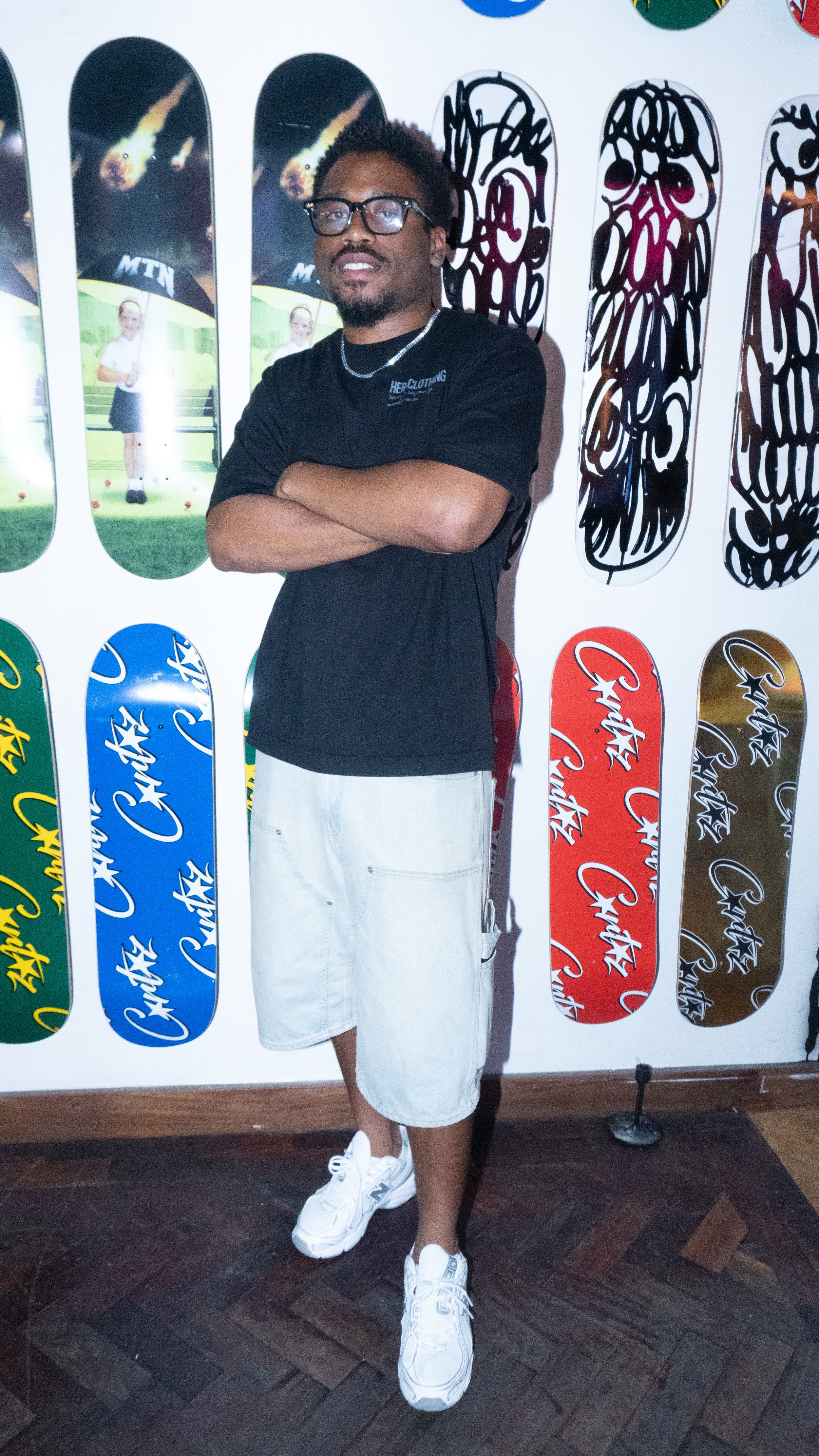 A young man with glasses and a beard standing with arms crossed in front of a wall decorated with skateboard graphics and colorful logos.