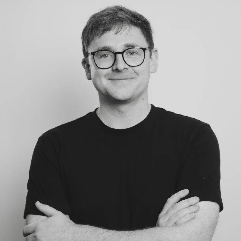 A young man with glasses, smiling, wearing a black t-shirt, with arms crossed, against a plain light background.